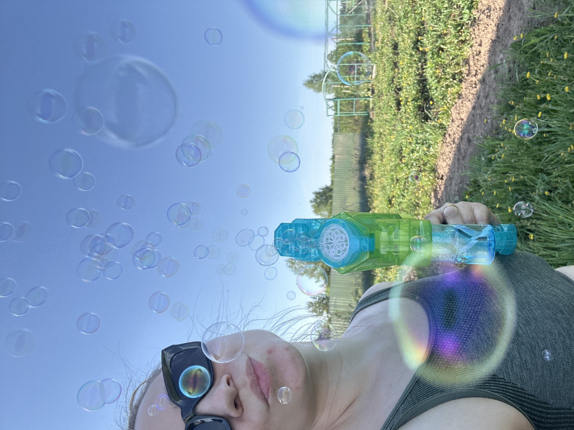 A woman is holding a blue and green bubble gun, blowing bubbles into the air. She is wearing sunglasses and a green tank top. The background includes a blue sky, green grass, a dirt path, and a green fence. The location cannot be determined with the given information.