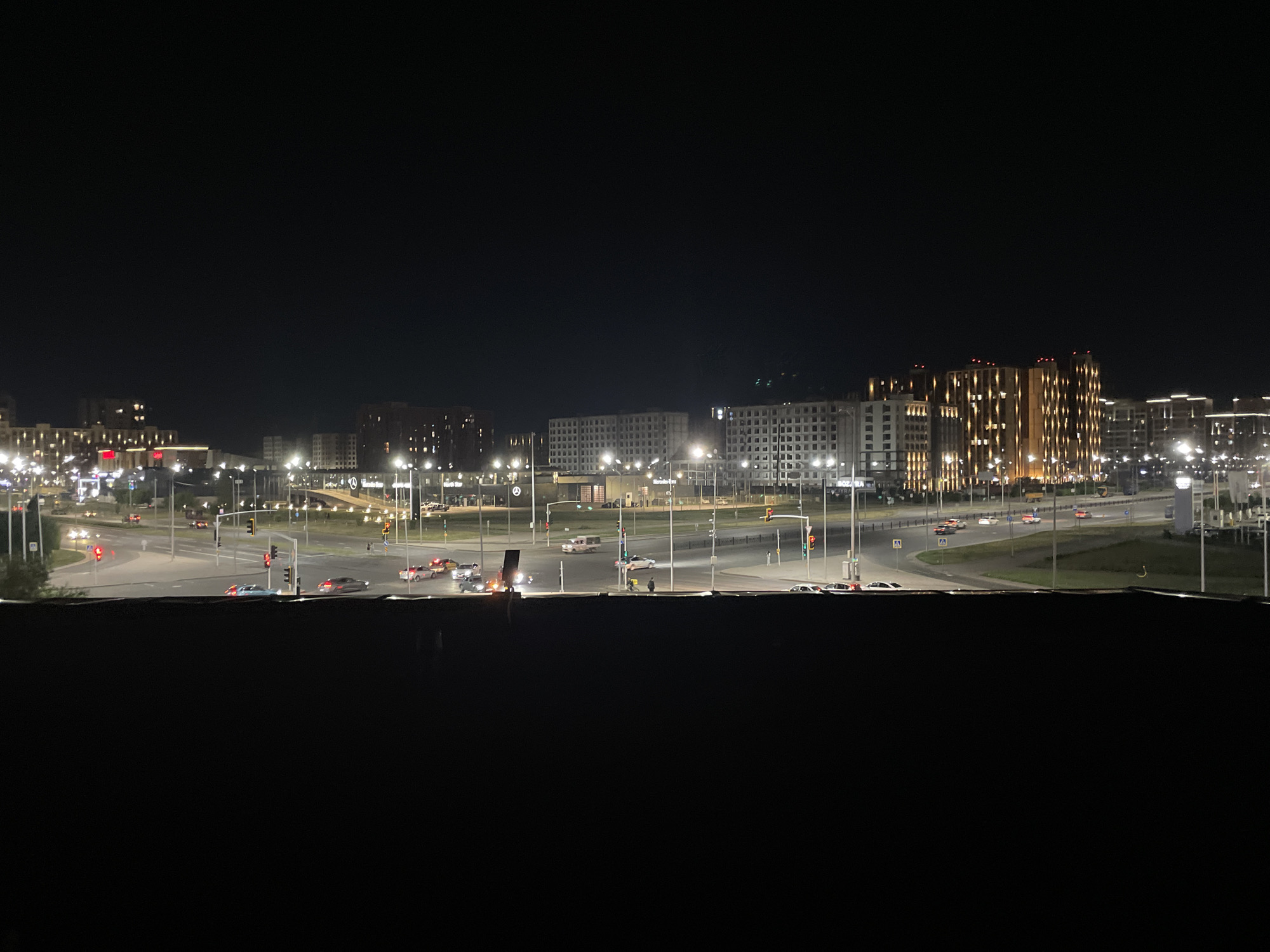 This is a nighttime panoramic view of a modern urban area in Astana, Kazakhstan. The foreground is dominated by a dark, out-of-focus area, likely a railing or window sill. Beyond this, the middle ground features a street intersection with several cars, some with their headlights on, navigating the roads. Traffic lights are visible and illuminated. In the background, multiple large, modern buildings are illuminated, some with intricate, glowing patterns on their facades, suggesting commercial or residential complexes. Streetlights line the roads and walkways, casting a warm glow. The sky is dark, indicating nighttime. The overall impression is of a well-developed and active city district at night.