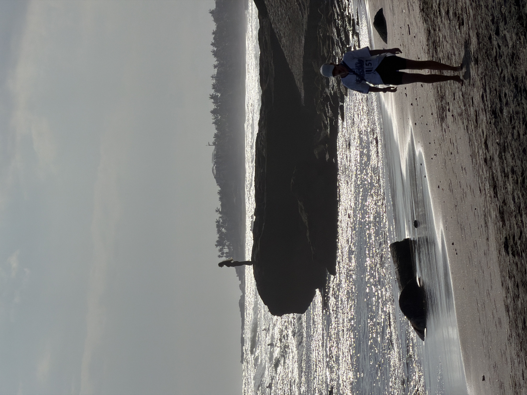 The image depicts a serene beach scene, likely in North Kuta, Indonesia, characterized by a silhouetted figure standing on a dark rock formation in the mid-ground. The ocean is visible with shimmering sunlight reflecting off the waves. In the foreground, another figure, a person wearing a cap, t-shirt, and shorts, walks along the sandy shore. Palm trees line the distant horizon, suggesting a tropical coastal environment. The sky is overcast, with diffused light creating a soft, atmospheric effect. The composition emphasizes the natural beauty of the coastline with the human presence as small elements within the grand landscape.