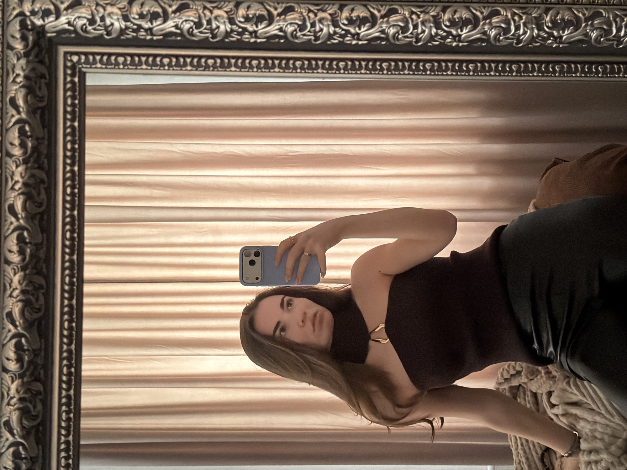 A person is taking a selfie in a mirror. The mirror has an ornate, antique-style frame. The background reflected in the mirror consists of horizontal blinds. The person is holding a light blue smartphone. The person is wearing a dark colored top and dark pants or leggings. The location is likely indoors. There is no information to suggest that the photo was taken in Moscow, Russia.