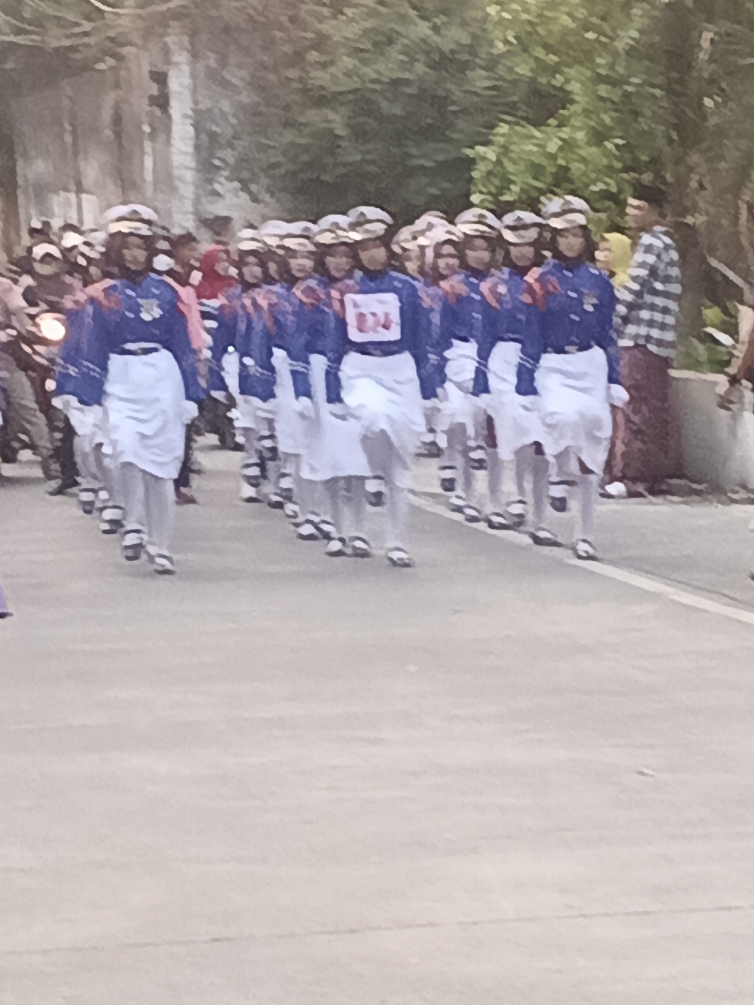 In Bogem Pinggir, Indonesia, a marching band comprised of individuals wearing blue uniforms with white skirts and white hats is parading on a paved road. The band members are positioned in a formation, with some individuals carrying white objects or flags. In the background, a person wearing a plaid shirt and a red sarong is visible, along with trees and a building. A motorcycle is also present on the left side of the image.