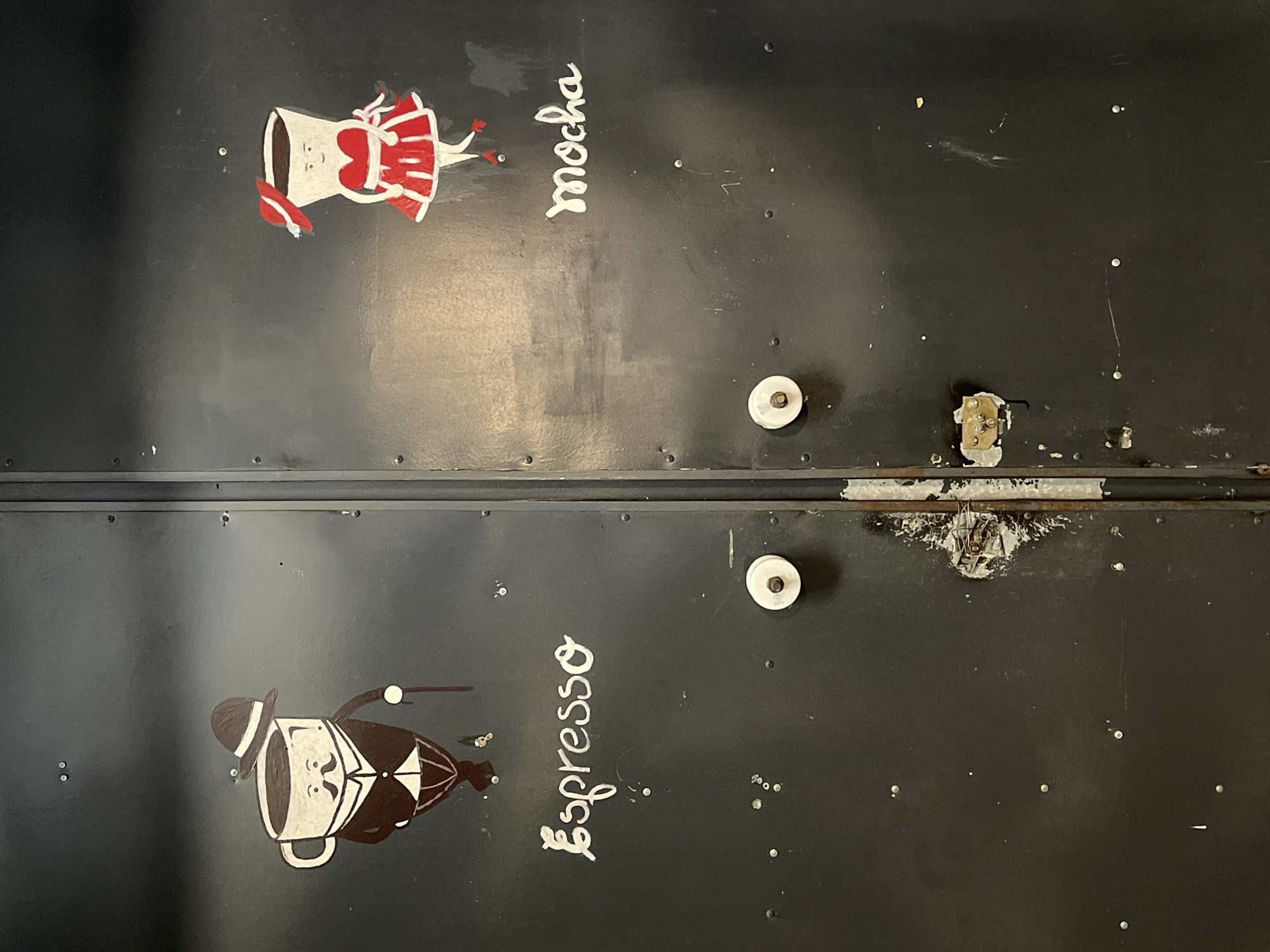 The image shows a pair of black doors with drawings and lettering. The upper door features a cartoon-like drawing of a coffee cup wearing a red dress and a white apron. Next to it, the word "mocha" is written vertically in white. The lower door features a cartoon-like drawing of a coffee cup wearing a black suit, hat, and holding a cane. Next to it, the word "Espresso" is written vertically in white. The doors are likely made of metal and have white knobs. A latch mechanism is visible between the doors, showing signs of wear. The location is Da Lat, Vietnam.