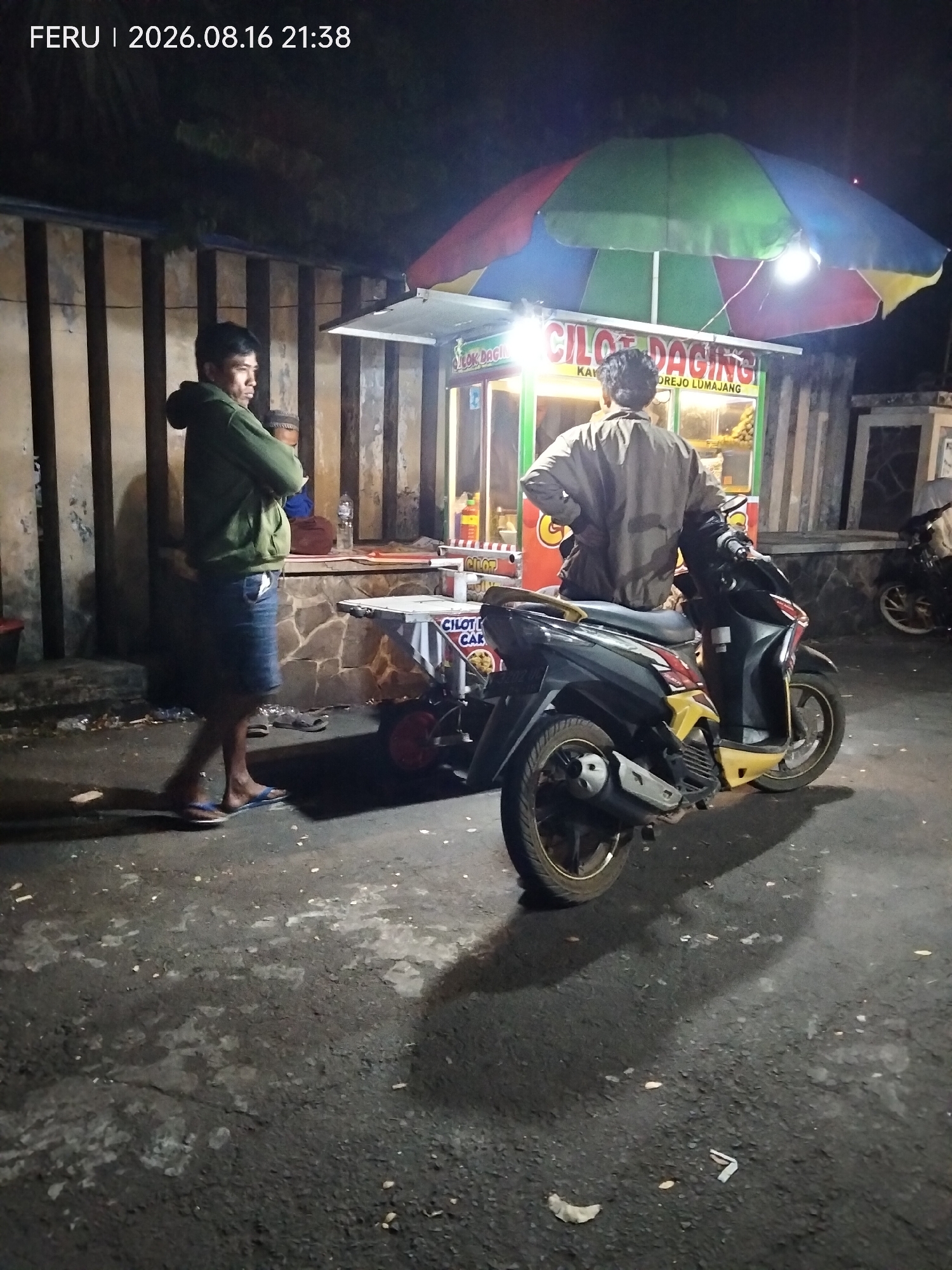 A wide shot captures an outdoor nighttime scene at a food stall in Rejoagung, Indonesia. The stall, illuminated by a brightly colored umbrella and overhead light, displays signage reading "CILOT DAGING". A man, wearing a green hooded jacket and denim shorts, stands with his arms crossed, facing away from the viewer and towards the stall. Another man, wearing a tan jacket, is seated on a black and yellow scooter parked in front of the stall, also facing away. The scooter's shadow stretches across the dark, asphalt ground, which is littered with debris. A second scooter is partially visible further back on the right. The background features a wall constructed of vertical wooden planks and rough stone.
