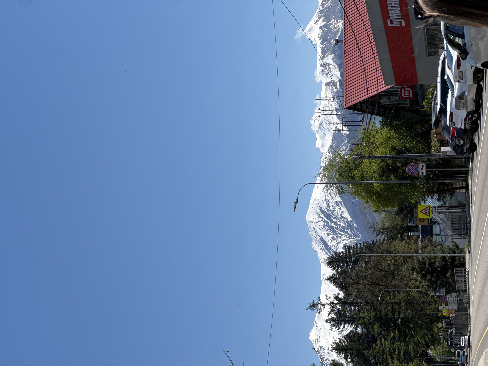 The image depicts a street scene in Krasnaya Polyana, Russia, with a clear blue sky dominating the left half of the frame. Towering snow-capped mountains form the backdrop. On the right side of the frame, there's a building with a red and grey facade, displaying a large logo. Adjacent to the building, a row of parked cars is visible. Street infrastructure, including lampposts and road signs, lines the roadside. Trees are interspersed along the street, adding greenery to the scene. The environment is urban yet set against a mountainous landscape.