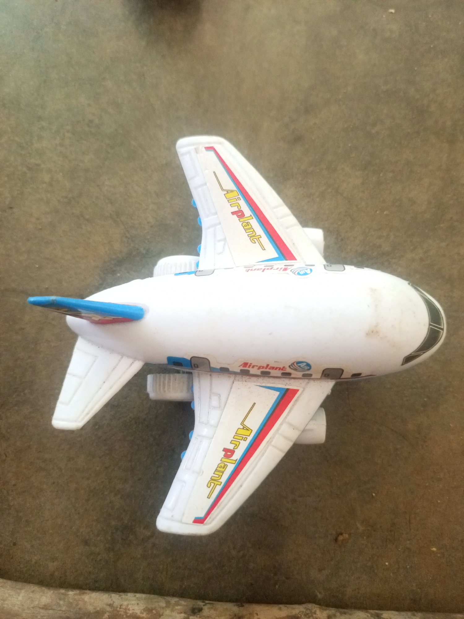 A white toy airplane is lying on a rough, textured surface that appears to be dirt or coarse sand. The airplane has blue and red stripes along its wings and fuselage, with the word "Airplane" printed in yellow on the wings. It has small blue accents on the engines and wingtips. The overall impression is of a child's toy that has been played with outdoors. The provided context mentioning "La Tagua, Venezuela" is not visually supported by the image itself.