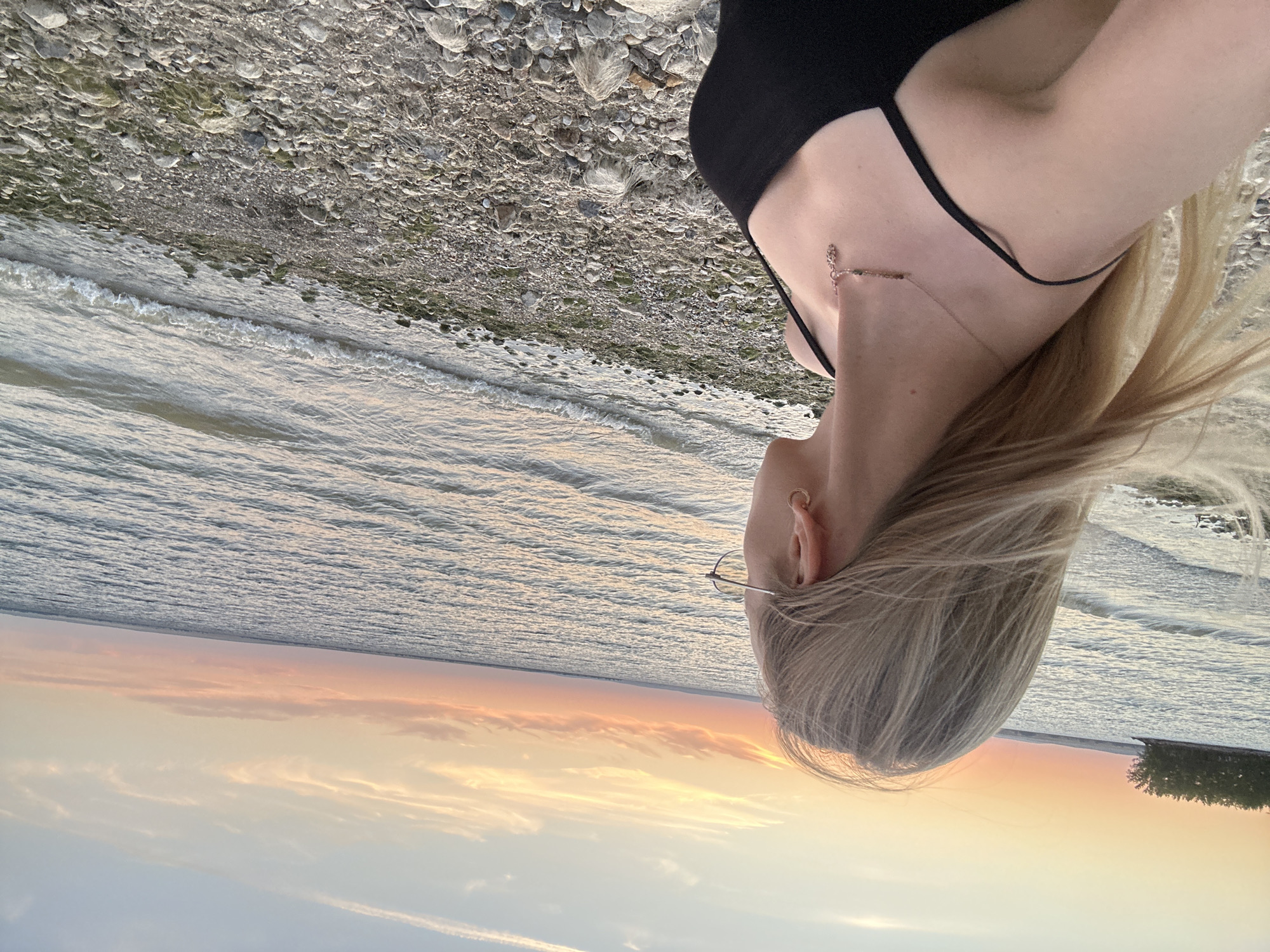 An over-the-shoulder medium shot captures a young woman from the chest up, looking out at a body of water during sunset. She has long, blonde hair that is blowing in the wind and wears glasses, a black tank top, and a delicate necklace. The background features a vast expanse of water with gentle waves and a sky filled with soft orange and pink clouds, indicating a sunset. The shoreline is covered in pebbles and some sparse vegetation. The scene is set in Energetik, Russia.
