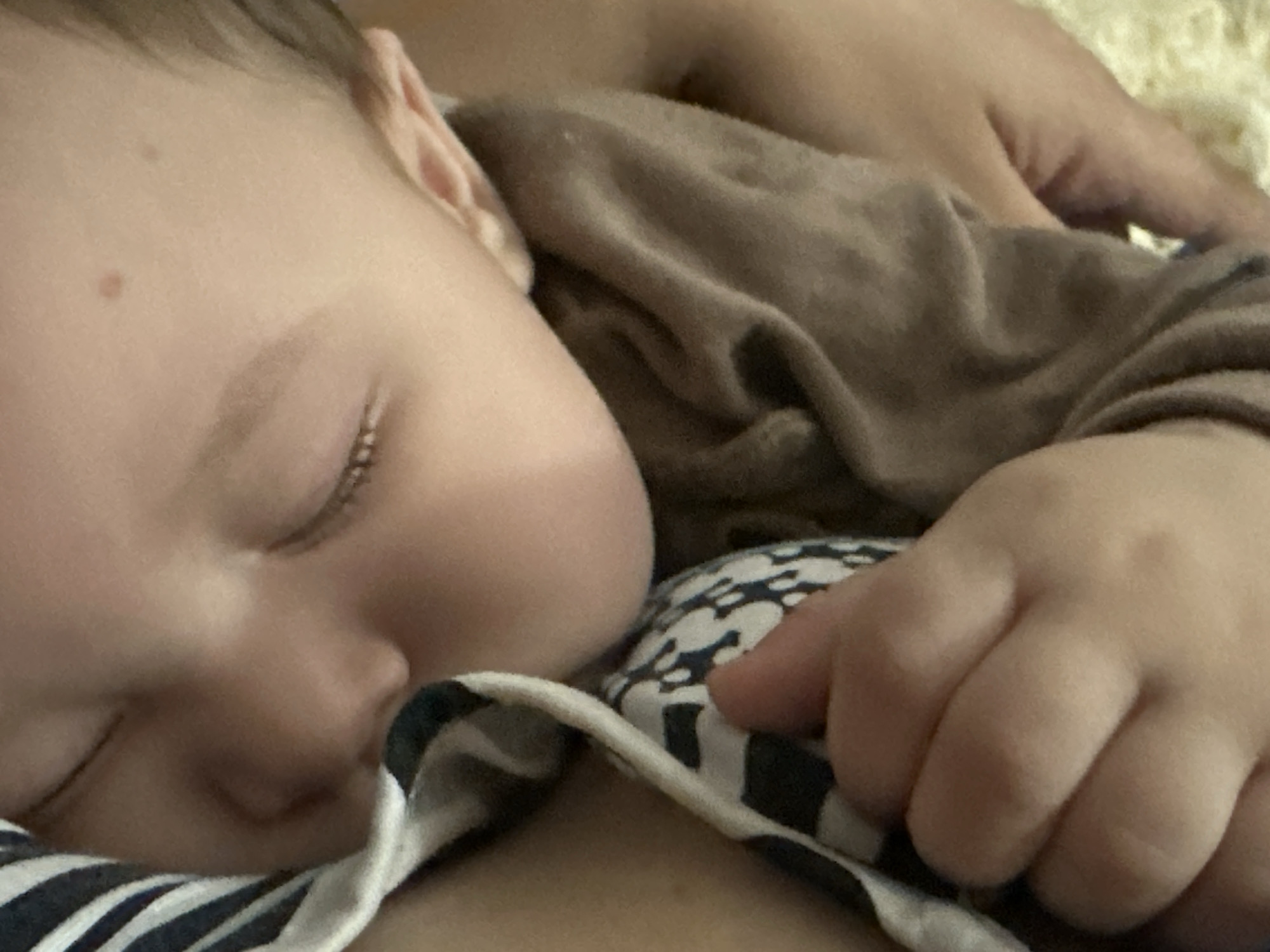 A close-up, intimate image captures a baby sleeping peacefully, nestled against an unseen adult. The baby is positioned on its side, facing to the left of the frame, with eyes closed and a serene expression. Its left cheek and nose are gently pressed against a black and white striped fabric, which appears to be soft and possibly part of a blanket or clothing. The baby is wearing a plain brown long-sleeved top, showing some wrinkles and folds in the fabric.
Visible details of the baby include smooth, fair skin, with delicate eyelashes resting on the cheek. Two small, reddish-brown marks are discernible on the baby's forehead. A tiny, chubby hand with curled fingers is resting on the striped fabric, contributing to the sense of a deep, comfortable sleep. In the upper right corner, an adult's hand is visible, gently resting on the brown fabric of the baby's shirt, suggesting a comforting presence. Further in the background, a soft, light-colored, possibly fuzzy textile is visible.
The scene conveys a strong sense of calm, intimacy, and security. The lighting is soft and subdued, implying an indoor setting, possibly during early morning, evening, or a dimly lit room, enhancing the peaceful atmosphere. No text is visible in the image.