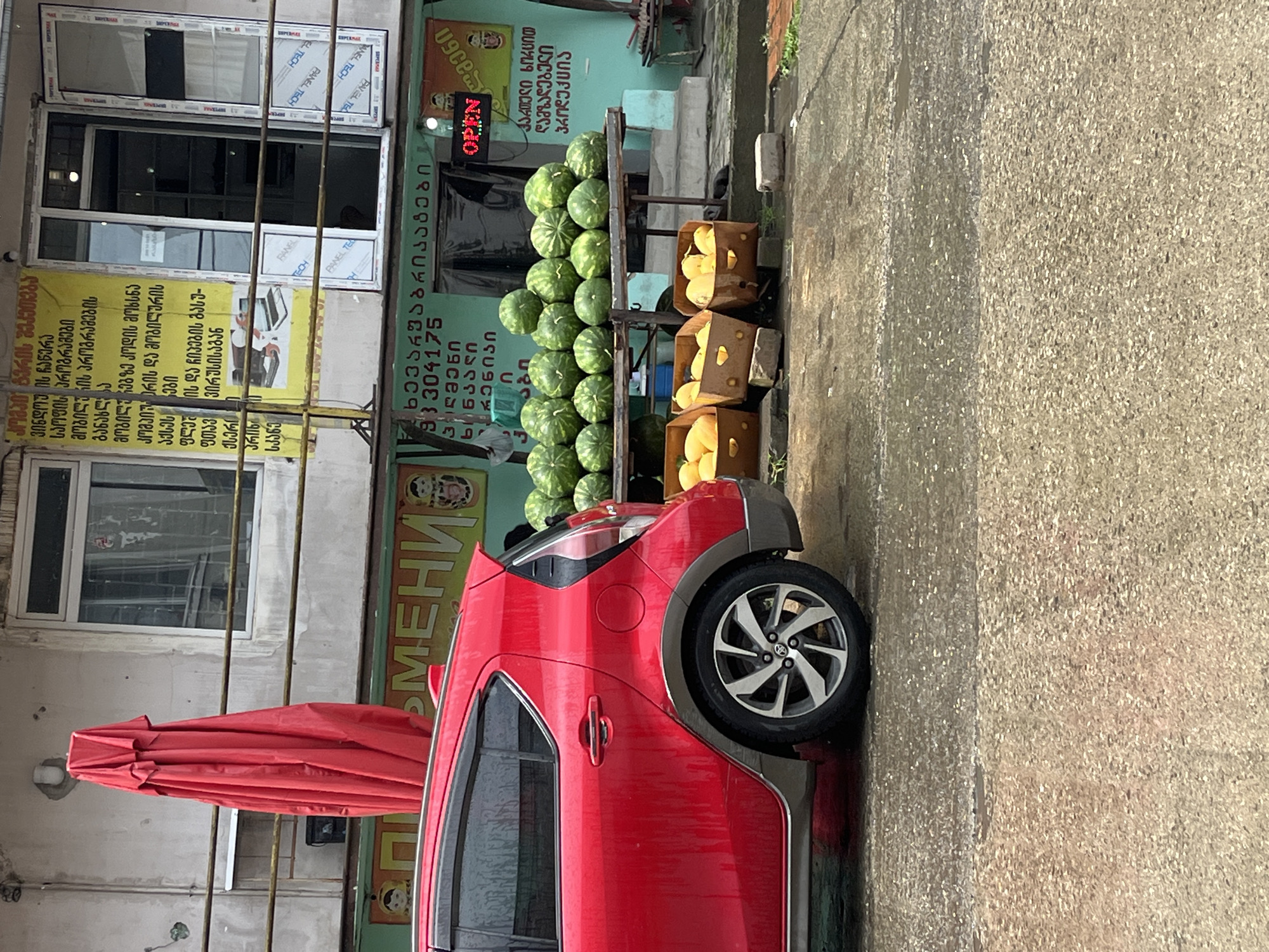 In Poti, Georgia, a red car is parked on a wet street in front of a storefront displaying watermelons and other produce. A large red umbrella is propped against the building. Signs in Georgian are visible on the building, including one that reads "OPEN" in red lettering. The scene appears to be an outdoor market or roadside stand.