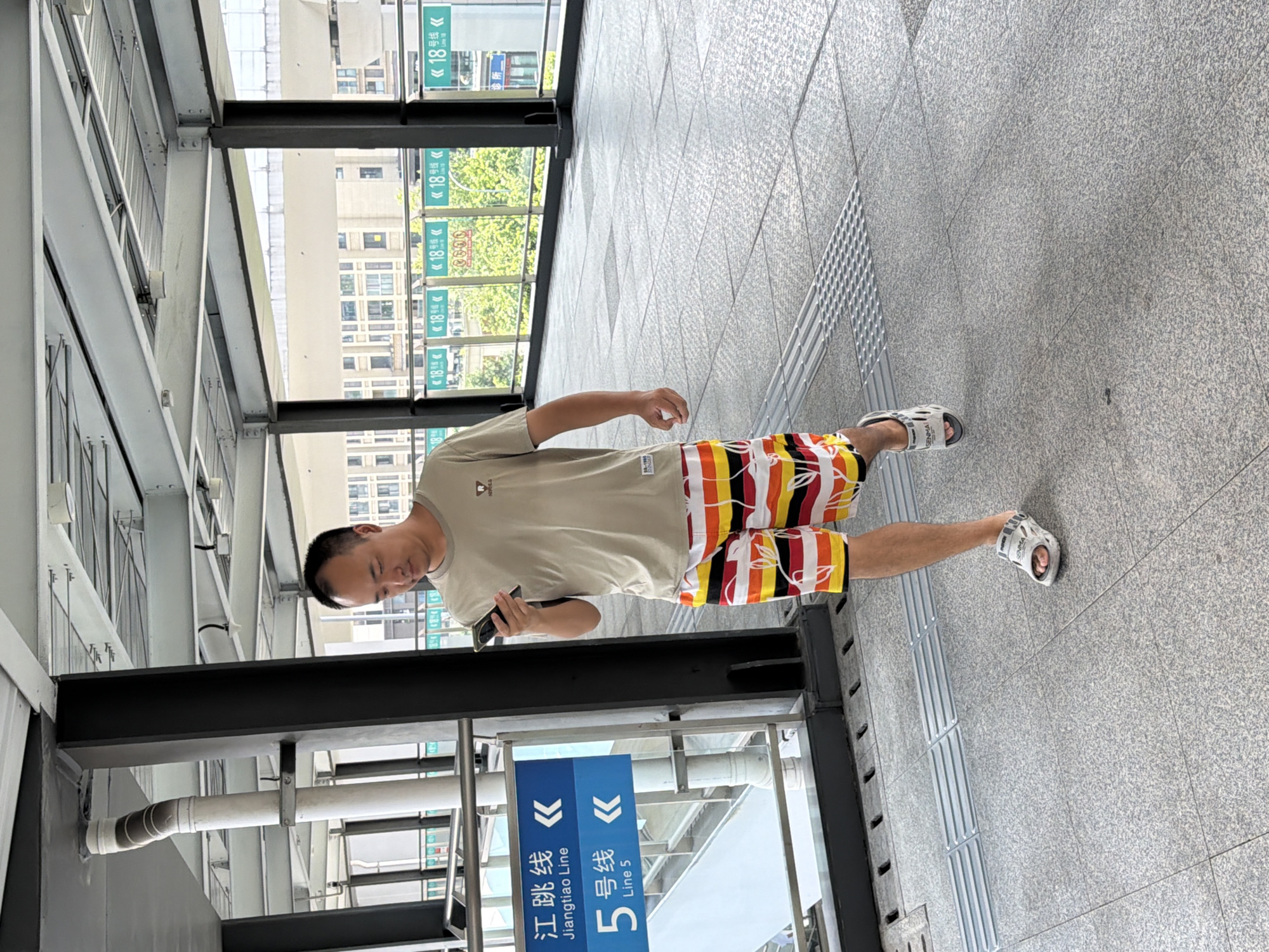 A man is walking on a pedestrian overpass in Tiaodeng, China, while looking at his phone. He is wearing a light-colored t-shirt, brightly colored striped shorts with a floral pattern, and white sandals. The overpass has a tiled floor with tactile paving and metal railings. In the background, there are signs indicating train lines and buildings. The man is positioned in the center of the frame, walking towards the viewer's right.