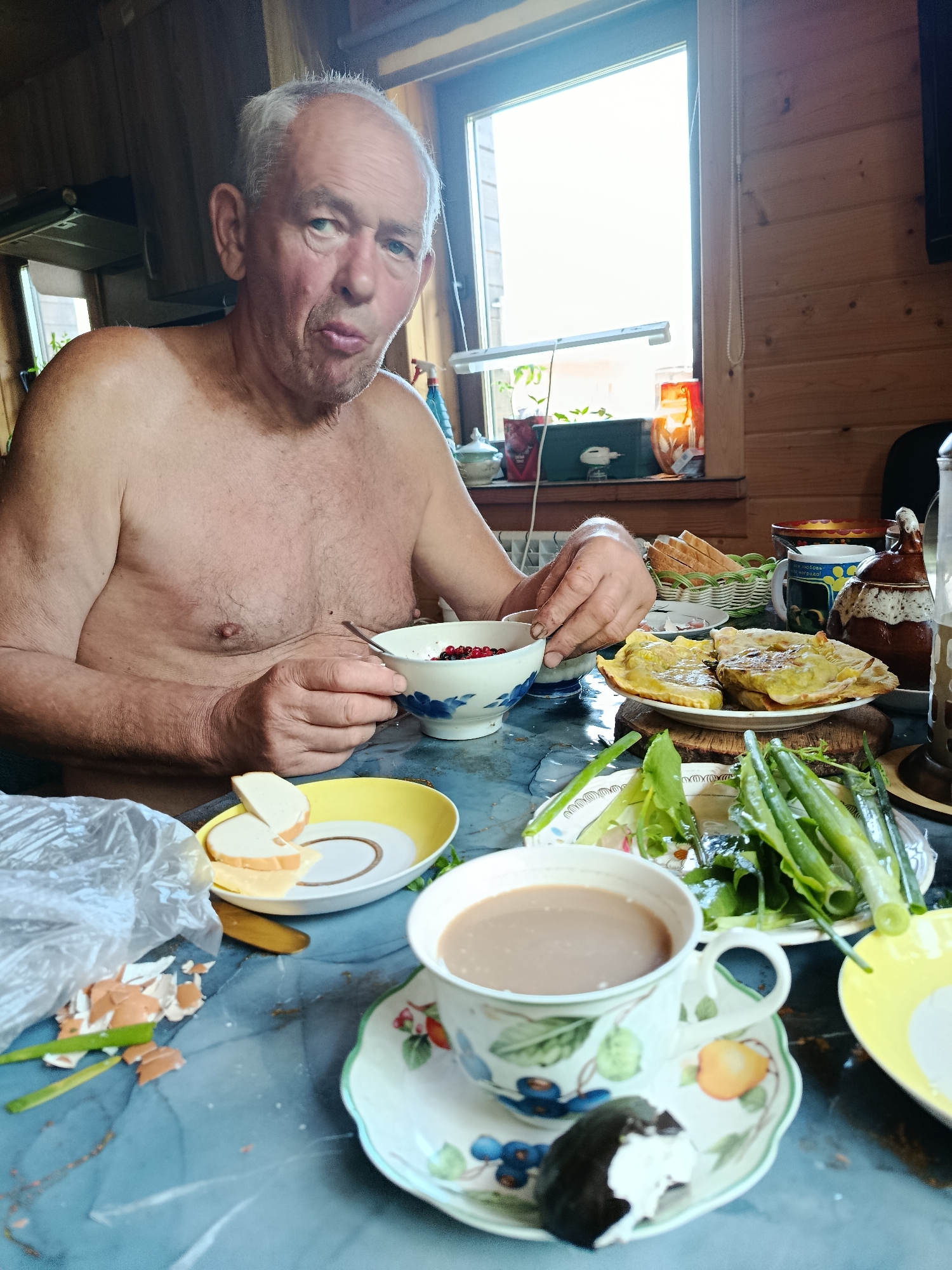 An elderly Caucasian male, shirtless and with thinning grey hair, is seated at a blue-patterned table in what appears to be a rustic indoor setting, likely a dining area or kitchen. He looks directly at the camera with a slightly pursed expression, holding a spoon in his right hand and dipping it into a white bowl containing red berries, possibly cranberries or lingonberries. His left hand rests near the bowl.
The table is laden with a variety of food items. To the man's left, a yellow plate holds two slices of white bread and a slice of cheese. Adjacent to this, a clear plastic bag is open, containing broken brown eggshells and green onion trimmings. To the man's right, a white plate with two golden-brown, folded pastries, possibly savory pies, rests on a wooden trivet. Further right, another plate holds green leafy vegetables and green onions. In the foreground on the right, a white teacup with a floral design (blueberries, oranges, leaves) sits on a matching saucer, containing a light brown beverage. A partially eaten dark pastry with a white filling is also on the saucer. In the background on the table, a small woven basket contains slices of bread, a ceramic mug with a handle and Cyrillic text, and a dark brown ornate teapot or pitcher.
The background consists of light-colored wooden plank walls, suggesting a wooden cabin or house. A large window on the right side provides bright daylight, washing out the external view. On the windowsill or a shelf below the window, several small potted plants or seedlings, a red-orange glass object, and a small white appliance are visible. A string of white beads hangs vertically by the window frame. To the left of the man, darker wooden cabinetry and what appears to be part of an exhaust fan or range hood are visible. A horizontal white strip light is mounted near the window on the left side. The scene is brightly illuminated by natural light. The location is Unknown, Unknown.