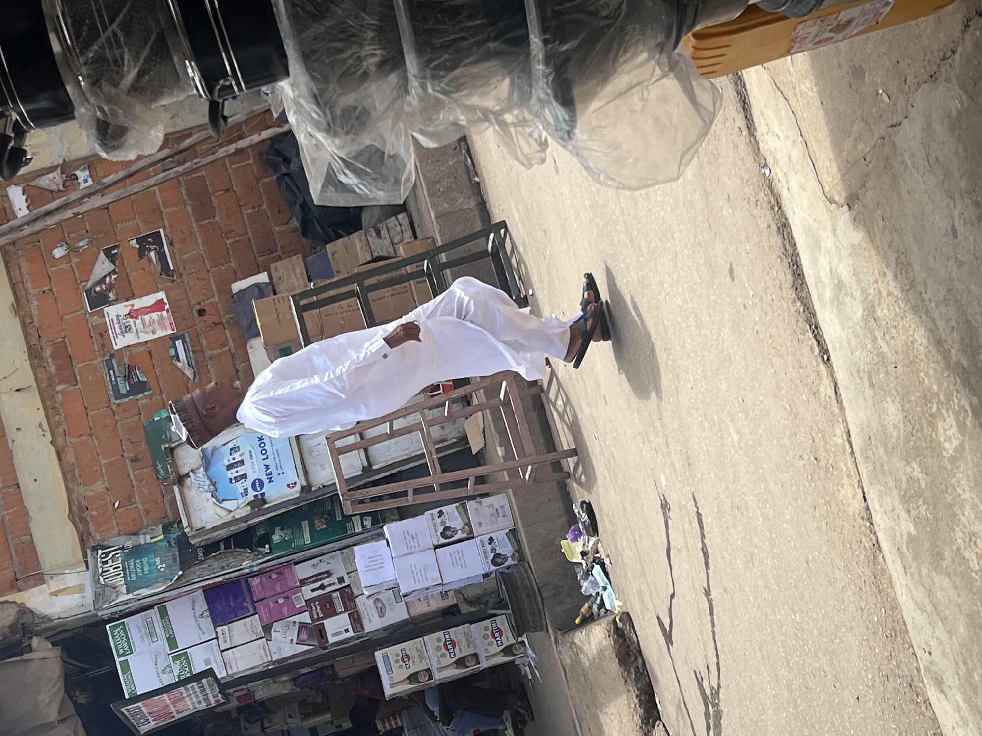 A man dressed in white is sleeping on a metal rack on a paved street in what appears to be a commercial district of Abuja, Nigeria. He wears sandals and is surrounded by shops stocked with packaged goods and drinks. The shops have signs advertising various brands, including William Lawson's, Martini, and New Look. The street is lined with a brick wall covered in posters. The overhead perspective may suggest the photo was taken from a higher position.