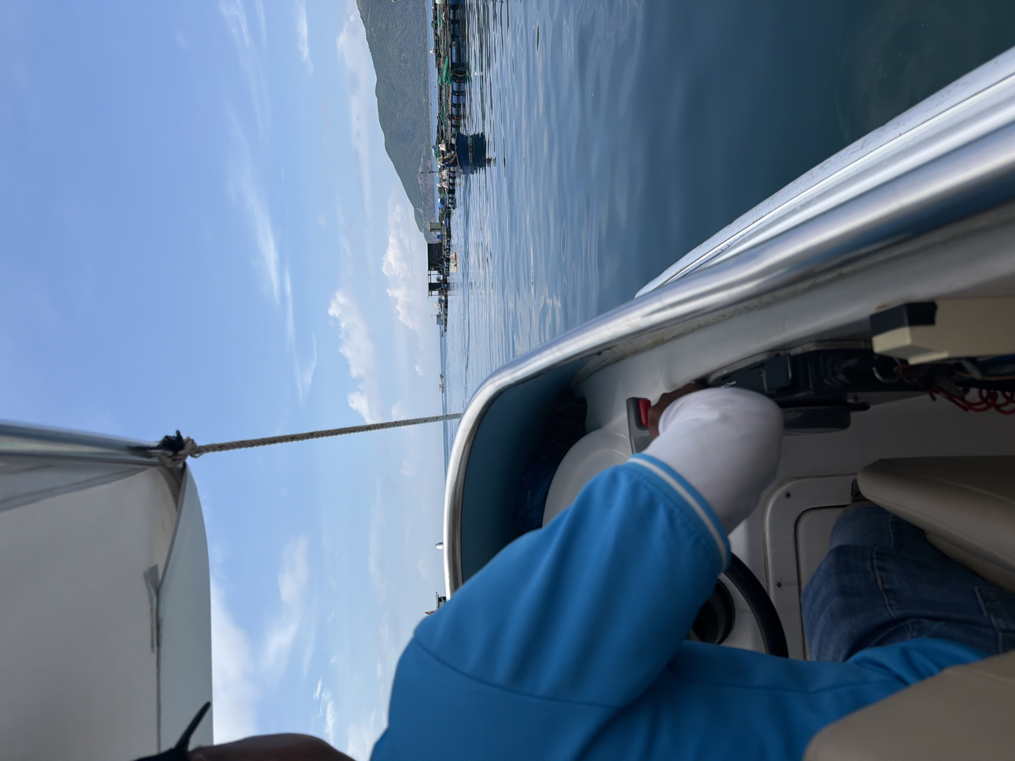 A person is sitting inside a boat, possibly steering. The person is wearing a blue shirt with white trim on the sleeves and blue jeans. The boat appears to be on the water, with a shoreline visible in the distance. The location is potentially Nam Nha Trang Ward, Vietnam. The weather looks clear with a few clouds in the sky.