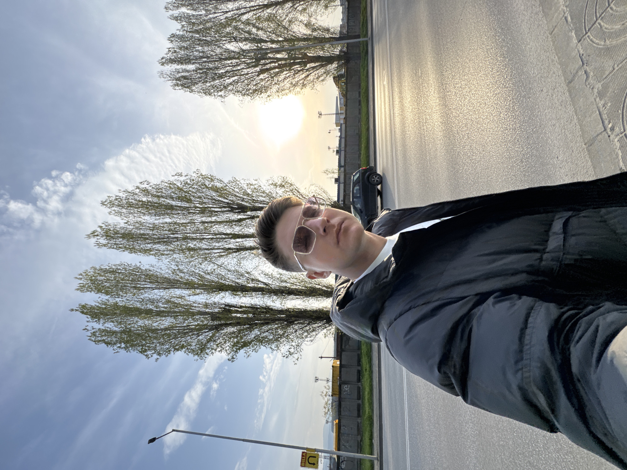 A medium shot shows a man standing on the sidewalk next to a road. The man is facing the camera, wearing sunglasses and a black jacket over a white shirt. He has short brown hair. To the left of the man, there are several bare trees and a street light. In the background, the sun is setting, and there is a row of container boxes. A car is parked on the side of the road. The setting is identified as Plovdiv, Bulgaria.