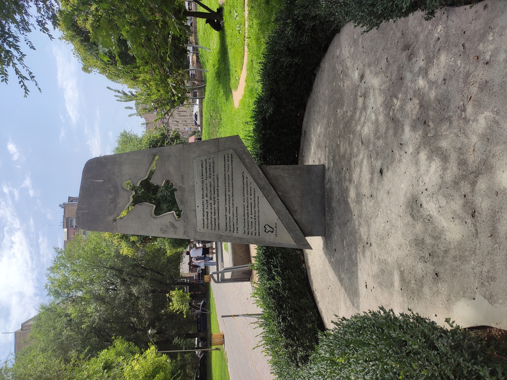 A stone monument stands in a park in Yerevan, Armenia. The monument is in the shape of an angled, abstract slab with a silhouette of a person in a dynamic pose cut out of it. Text in Armenian and English is inscribed on the monument, indicating it commemorates the "Yerevan Skatepark" initiated and built by the "Union of Armenians of Ukraine" in memory of Grigor Vllun Shatvoryan and donated to the city of Yerevan. The date "15.11.2021" is also visible.
The monument is situated on a circular concrete pad, surrounded by well-maintained green hedges. Behind the monument, a paved walkway curves into the distance, lined with trees and a lamppost. In the background, buildings and more trees are visible under a partly cloudy sky. A few people are seen walking on the paved path near the monument. The overall scene depicts a public park with a commemorative structure, suggesting a place for recreation and remembrance.