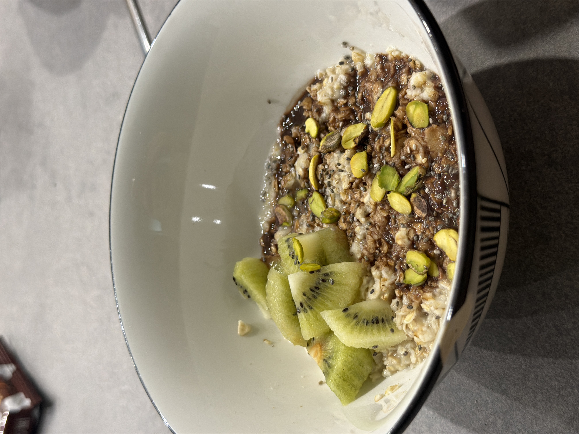 A close-up, overhead shot shows a bowl of oatmeal topped with kiwi slices, pistachios, and chia seeds. The oatmeal has a brown swirl of what appears to be chocolate sauce. The bowl is white with a black rim and a black geometric pattern on the outside. It sits on a gray, speckled surface. In the upper left corner, a blurred package of cookies is visible. The lighting suggests indoor illumination. No specific location in Vladivostok, Russia, can be determined from the image.