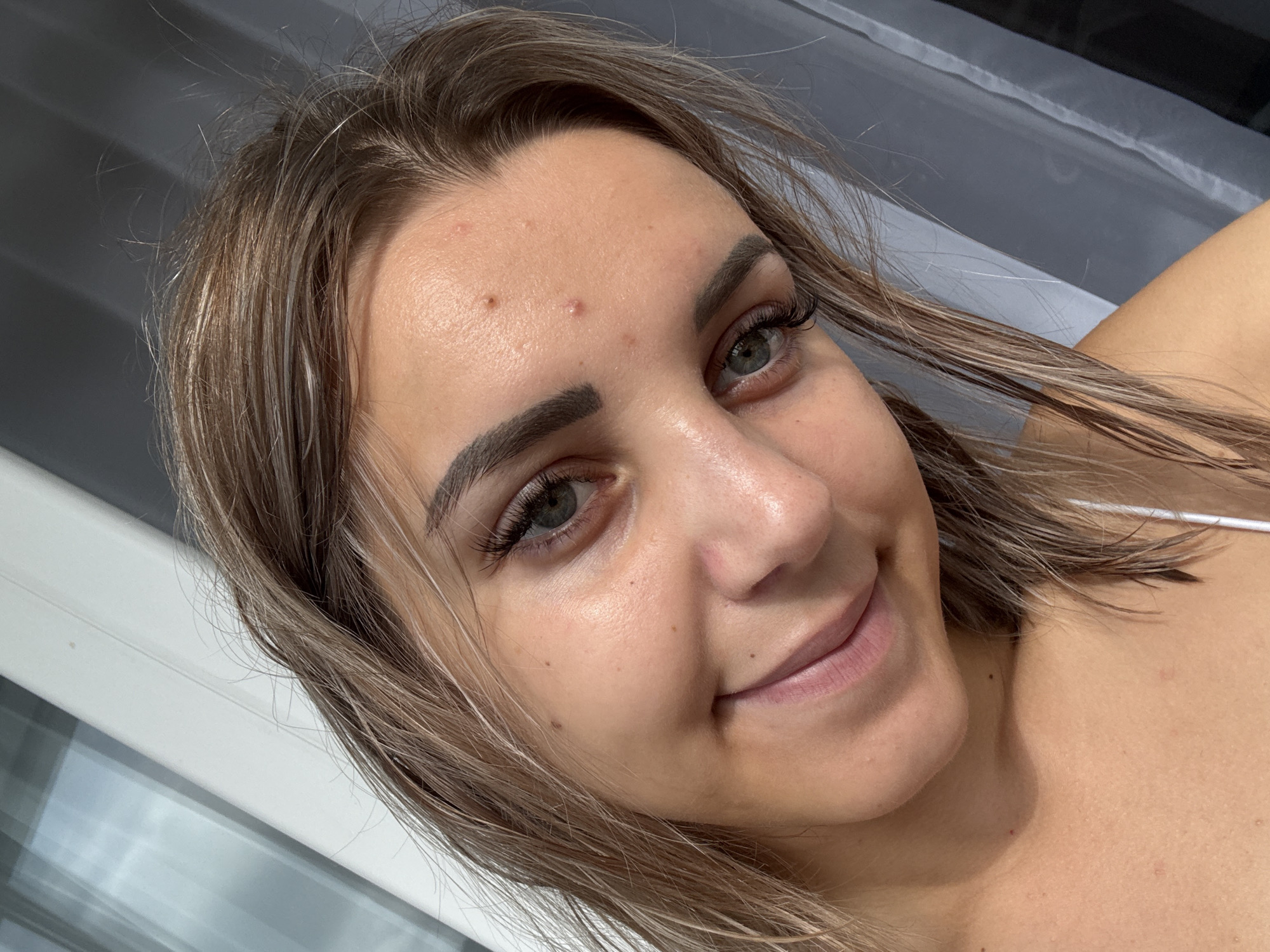 The image is a close-up selfie of a young woman. She has fair skin, light brown hair with blonde highlights, and green eyes. She is wearing minimal makeup and has several pimples on her forehead. She is smiling gently, and her head is tilted slightly to her left. The background appears to be an indoor setting with blinds and curtains visible. There is no discernible information in the image to suggest a location in Kholmsk, Russia.