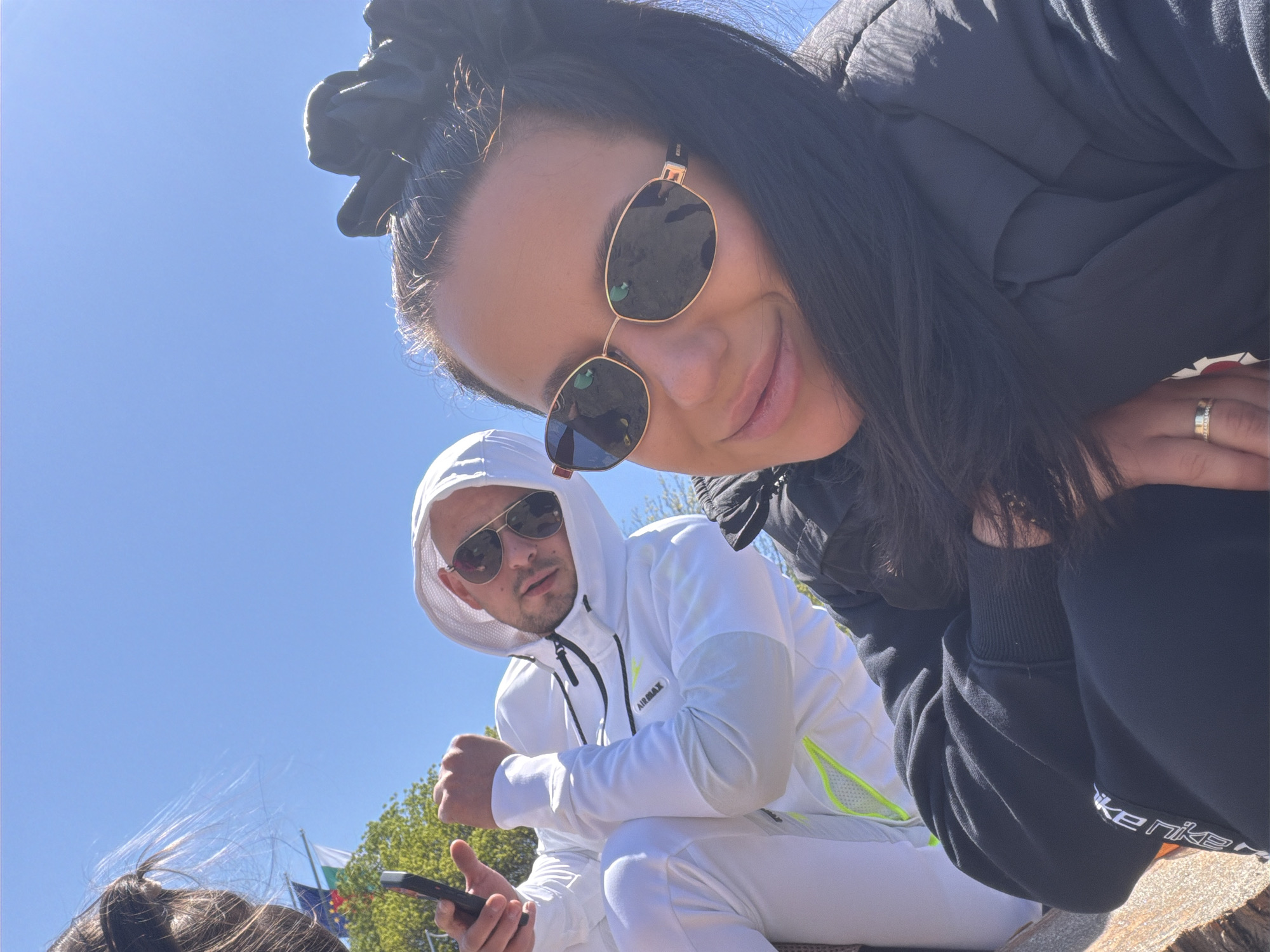 A low-angle shot shows a woman and a man outdoors. The woman, closer to the camera, wears dark sunglasses, a black jacket, and a hair scrunchie. She has dark hair. A gold ring is on her finger. The man, positioned behind and to the left of the woman, wears sunglasses and a white hooded tracksuit with yellow and black accents. He holds a dark-colored smartphone. Behind them, the sky is blue, and at the lower left, a small portion of hair and a flag are visible. The environment appears to be a public space in a city. "AIRMAX" is printed on the man's jacket, and "Nike" is printed on the woman's jacket.
