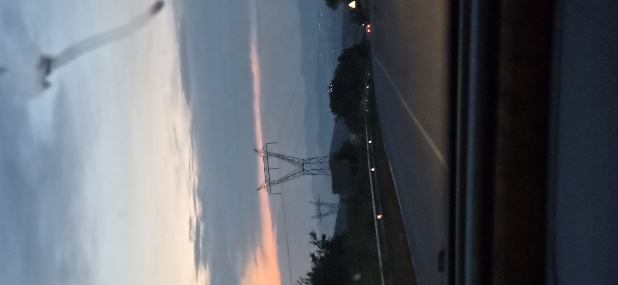 The image depicts a scene from a vehicle moving along a road at dusk. In the distance, a power line tower is visible against a sky with hints of orange and pink from the setting sun, juxtaposed with dark clouds. The road ahead curves gently to the right, bordered by a metal guardrail with reflective markers. Trees and hills form the backdrop, with distant city lights faintly visible on the horizon. The interior of the vehicle is partially visible in the foreground, showing a dark dashboard. The overall atmosphere is calm and suggestive of travel during twilight. The location is identified as being in Radilovo, Bulgaria.