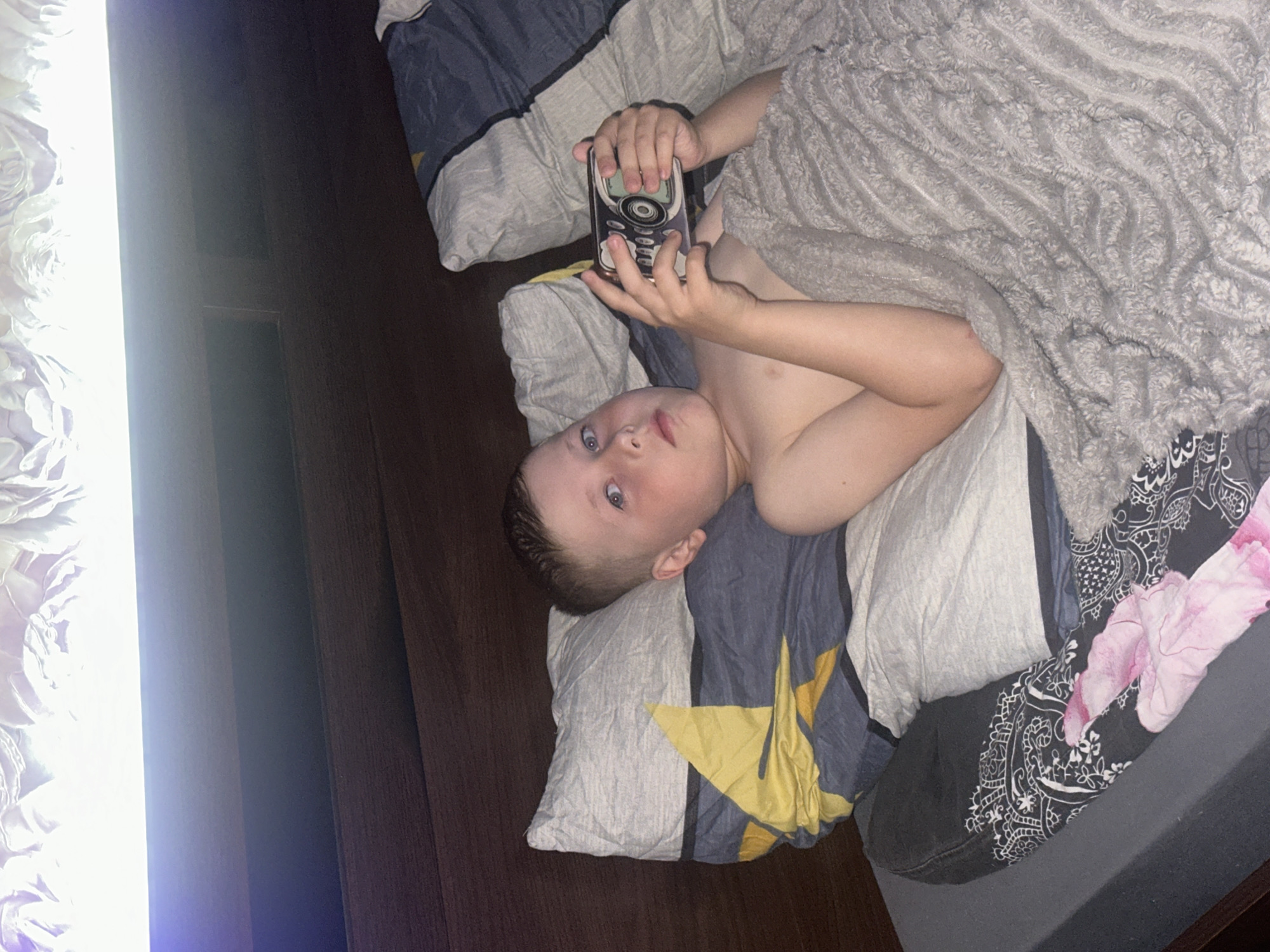 A young boy is lying in bed, holding a baby monitor. He is looking at the device with a curious expression. The bed is covered with pillows and blankets, one of which has a floral pattern. The room appears to be dimly lit, suggesting it is nighttime or early morning. The boy's face is visible, with blue eyes and light brown hair. He is wearing a tank top. A bright light source is visible on the left side of the image, casting a strong glow. The background shows a dark wooden surface, possibly a headboard or the wall. The overall scene evokes a sense of quiet domesticity.