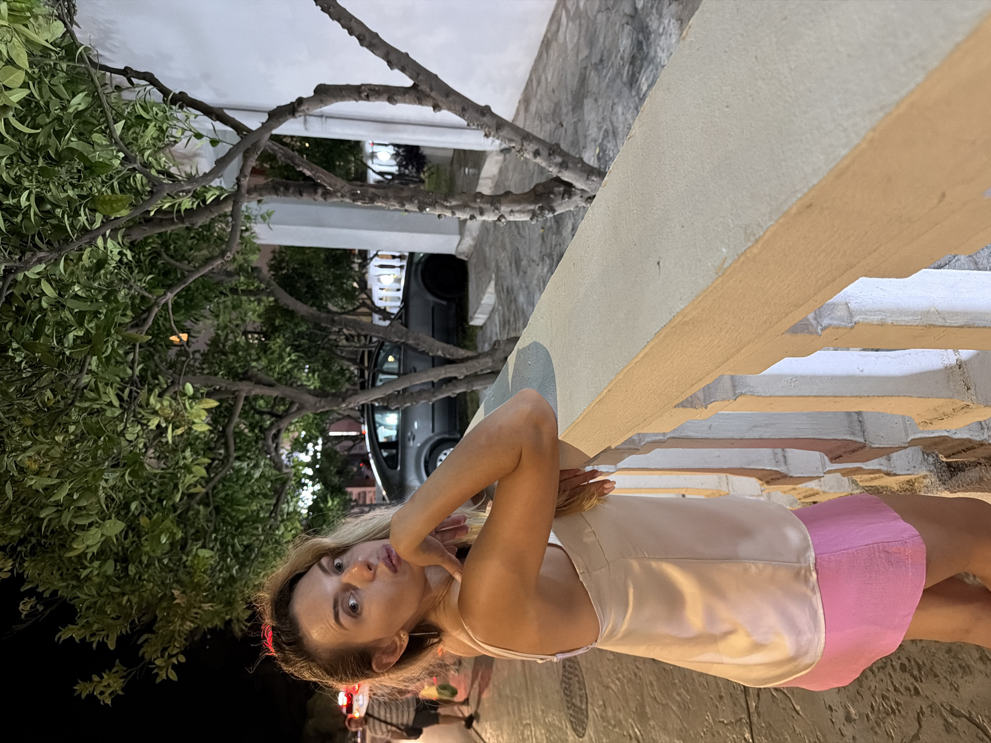 A woman with blonde hair, wearing a light pink camisole and a pink skirt, poses for a photo at night. She is leaning on a light-colored railing, with her right elbow on top of it and her left hand touching her chin. Behind her, trees with green leaves line a sidewalk, and a dark-colored car is parked on the street. The scene appears to be in Budva, Montenegro.