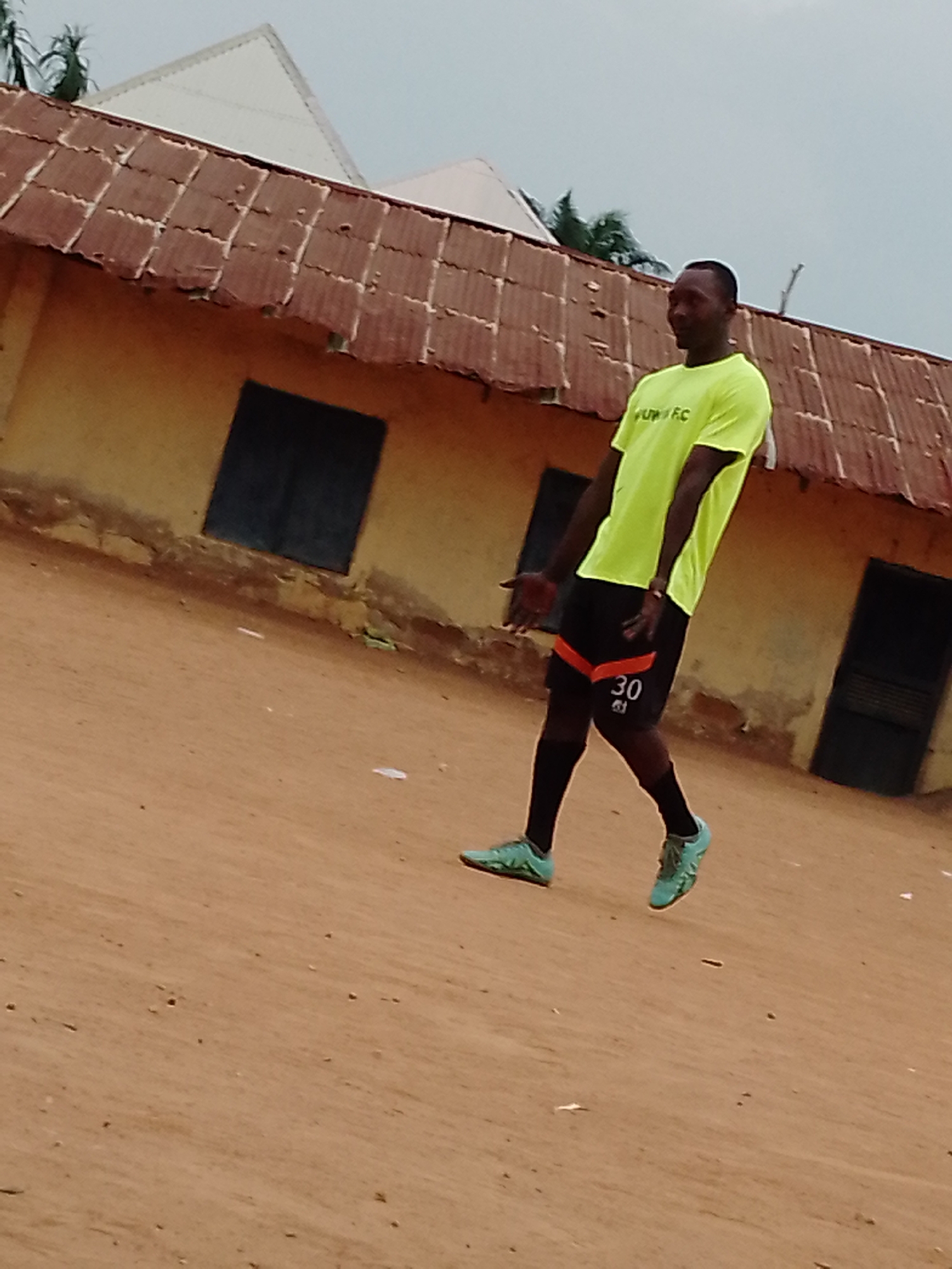 A man in a yellow shirt and black shorts is standing on a dirt surface in front of a yellow building with a rusty metal roof. He is wearing black socks and turquoise shoes. The building has two black doors and two black windows. The sky is overcast. The location is in an unknown city, unknown.