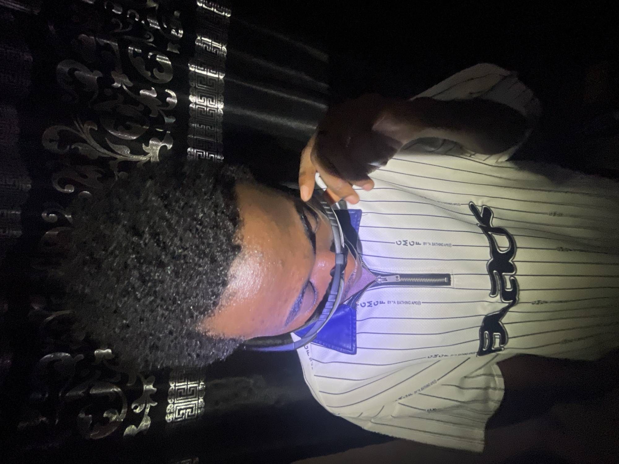 A person is lying down with their head on a decorated surface, possibly a bed. The person is wearing a white pinstripe shirt with a blue collar and a zipper. They also appear to be wearing headphones. The location is not discernible from the image; there's no information to suggest the location is Lokoja, Nigeria.