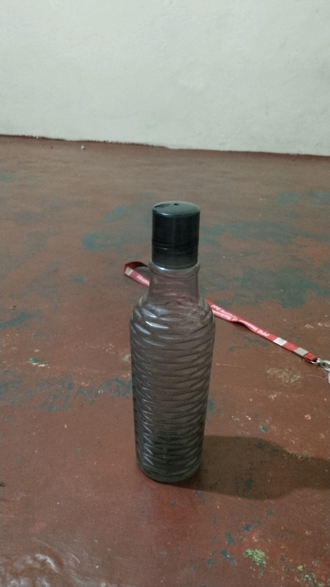 A plain grey bottle with a black cap is placed on a brown, speckled floor. A red and white lanyard is draped behind the bottle. The background consists of a white wall above a thin, dark line. The lighting suggests an indoor environment. The image's composition and subject matter do not provide any specific clues to pinpoint its location within Coimbatore, India.