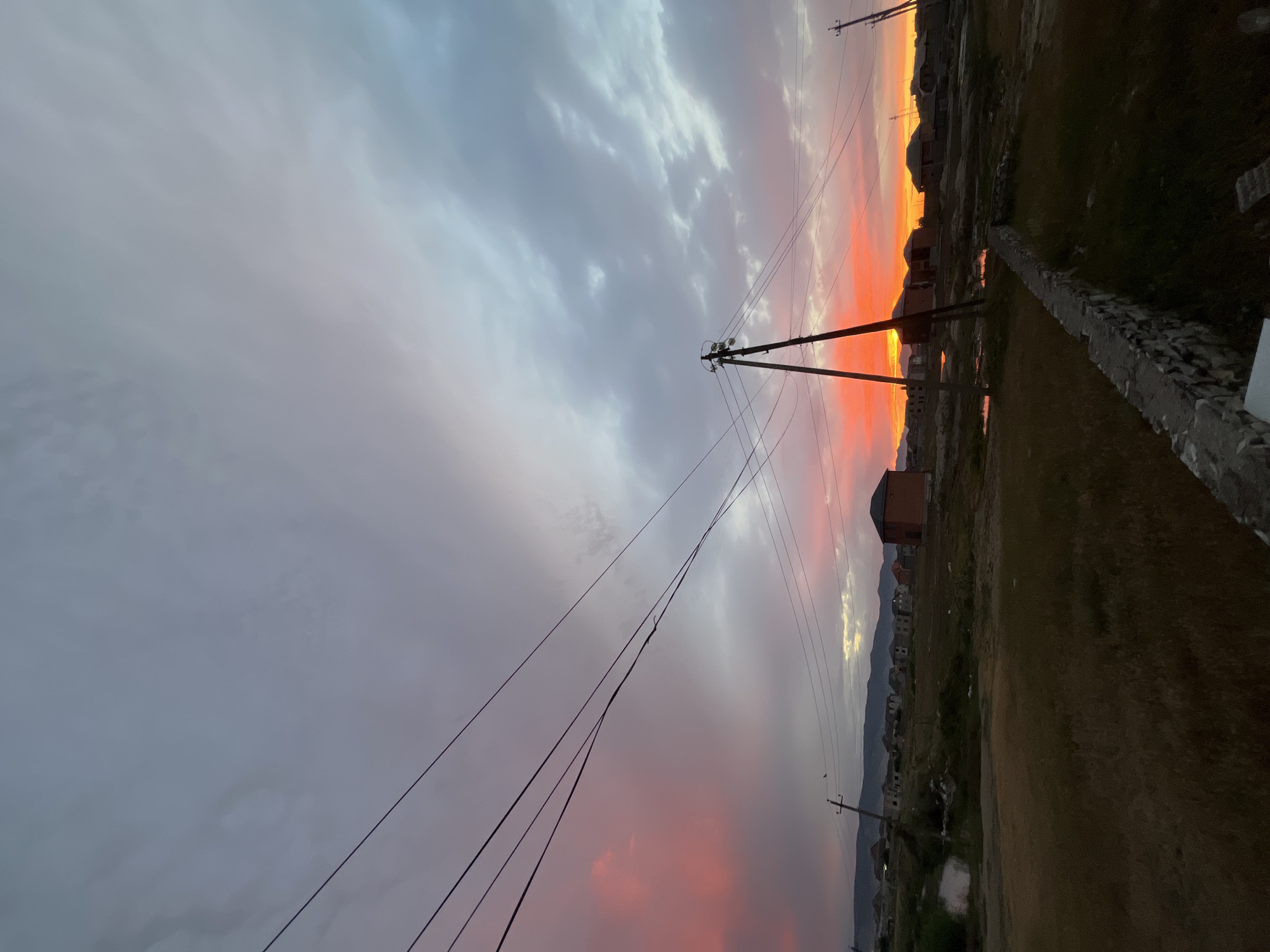 This image presents a serene and dramatic sunset over a developing landscape in Kaspiysk, Russia. The sky is the dominant feature, filled with a dynamic interplay of colors and clouds. Towards the top, the clouds are a soft grey and white, gradually giving way to intensely vivid oranges, reds, and yellows closer to the horizon, indicative of a spectacular dusk.
Several utility poles, likely made of wood or concrete, stand tall in the foreground and midground, their dark silhouettes starkly contrasting against the colorful sky. Numerous electrical wires crisscross the frame, adding a sense of human infrastructure interwoven with the natural beauty.
The ground below appears uneven and relatively barren, with patches of dry earth or sparse vegetation. A low, rough-hewn stone or concrete wall runs diagonally across the right side of the frame, leading towards the horizon. Scattered across the midground and horizon are various houses and buildings, mostly appearing as dark silhouettes. These structures suggest a semi-urban or rural settlement, possibly under development or situated on the outskirts of a town.
In the far distance, a distinct silhouette of mountains or hills stretches along the horizon line, providing a majestic natural backdrop to the scene. Small, reflective patches on the ground hint at the presence of puddles, subtly mirroring the sky's glowing hues.
The time of day is clearly sunset, imparting a warm and tranquil atmosphere. The weather appears partly cloudy, allowing for the striking color palette in the sky. No individuals or discernible text are visible in the image.
