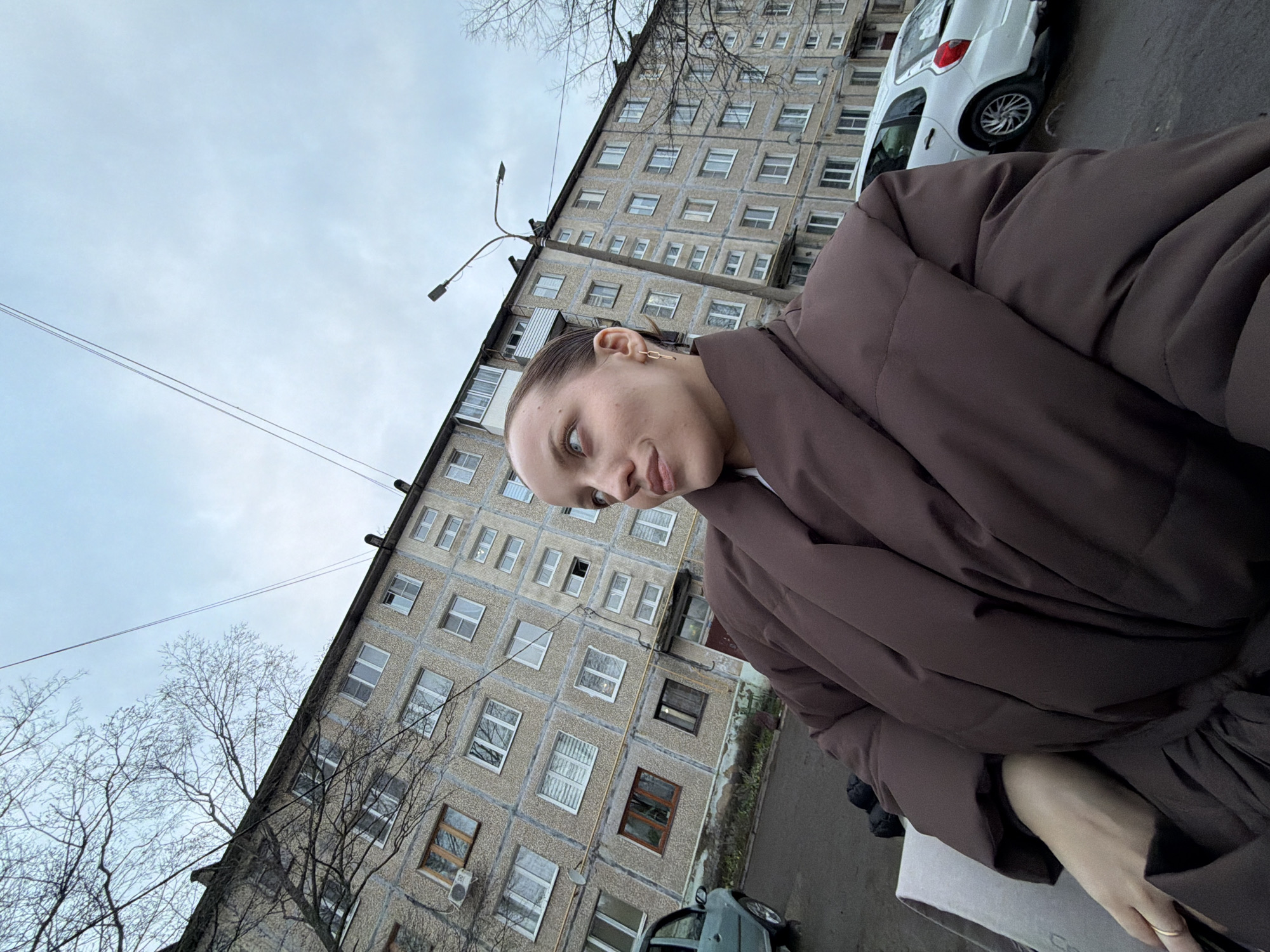 A tilted, low-angle selfie captures a young woman against a backdrop of a residential building under an overcast sky in Reshetnikovo, Russia. The woman, who appears to be in her twenties, has light skin, dark hair pulled back, and light eyes, directed towards the camera with a subtle smile. She is wearing a thick, dark brown puffer jacket and a single gold-colored chain earring is visible on her left ear. Her right arm and hand, adorned with a simple ring, are partially visible in the bottom right corner of the frame.
The dominant feature of the background is a large, multi-story concrete apartment block, characterized by rows of small, rectangular windows and visible panel construction, indicative of Soviet-era architecture. Bare tree branches extend into the left side of the frame, suggesting a cold season such as late autumn, winter, or early spring. The sky is a uniform light grey, indicating cloudy weather.
Other objects in the scene include a street light with multiple arms positioned above the building, and utility wires stretching across the upper portion of the sky. Two cars are partially visible on the ground level: a white car on the right and a smaller, darker car on the left. The setting is clearly an urban or suburban residential area. No readable text is discernible in the image.