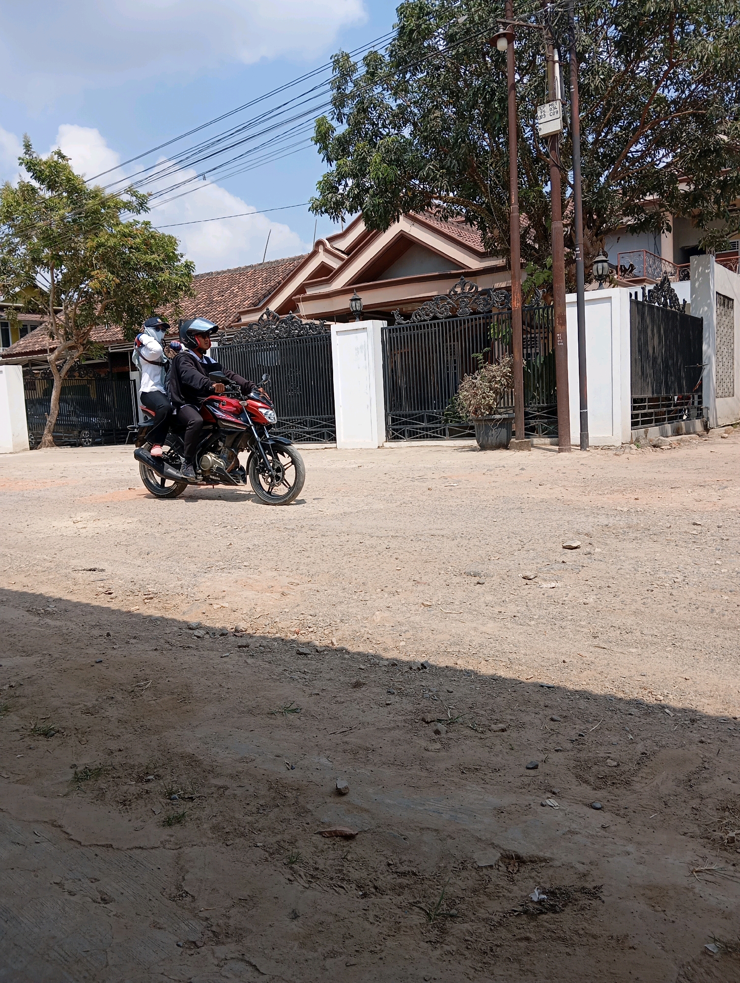 Two individuals are riding a red and black motorcycle on a dirt road under a clear sky. The person in front is wearing a black jacket and a helmet, while the person in the back is wearing a white shirt, black pants, and a hat, and appears to be holding onto the rider. They are passing a black metal fence in front of a house with a tile roof. The environment is rural or suburban, with trees and utility poles visible. The ground is covered in dirt and small stones, with some patches of sparse vegetation. The scene is illuminated by bright sunlight, casting shadows.