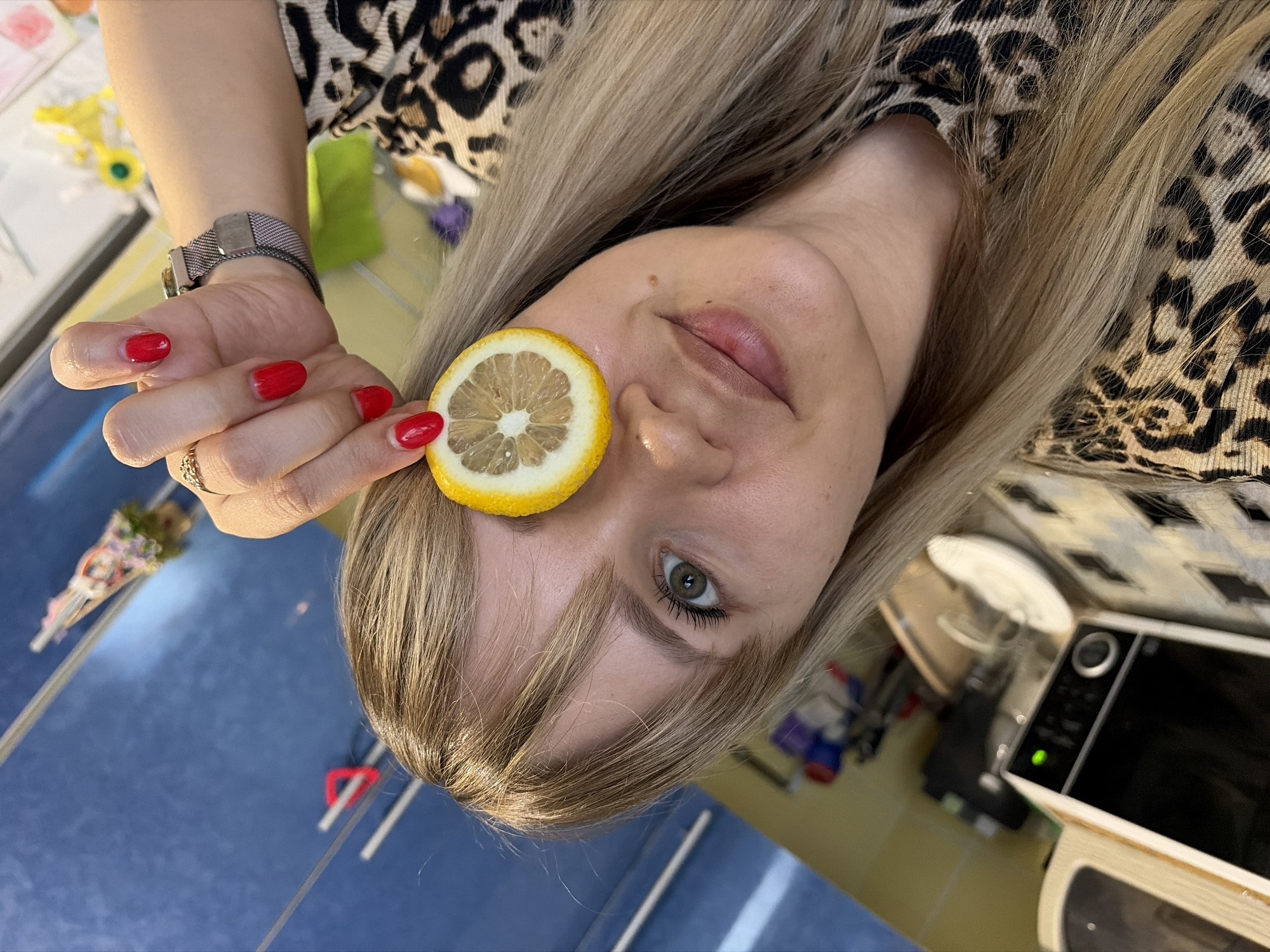 A woman with blonde hair is holding a slice of lemon on her forehead. She has red nail polish, a watch, and a ring on her left hand. She is wearing an animal print top. The background includes a blue surface, a microwave oven, and various other objects. The city and country of Chaikovsky, Russia, are not identifiable in this image.