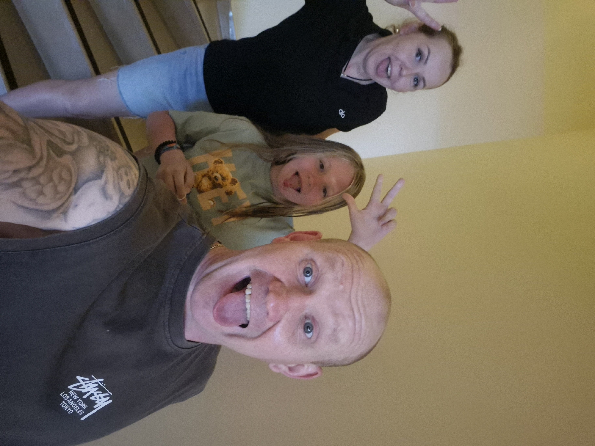 Three individuals pose for a photograph in what appears to be an indoor setting near Alanya, Turkey. A bald man wearing a gray Stussy t-shirt is in the foreground, sticking his tongue out. A girl with blond hair and a light green t-shirt with a bear design is positioned behind him, also sticking her tongue out and making a peace sign. Behind her, a woman with auburn hair and a black top is also making a peace sign and sticking her tongue out. The background includes a wall painted in tan or light yellow. The presence of stairs is evident in the upper left corner of the frame.