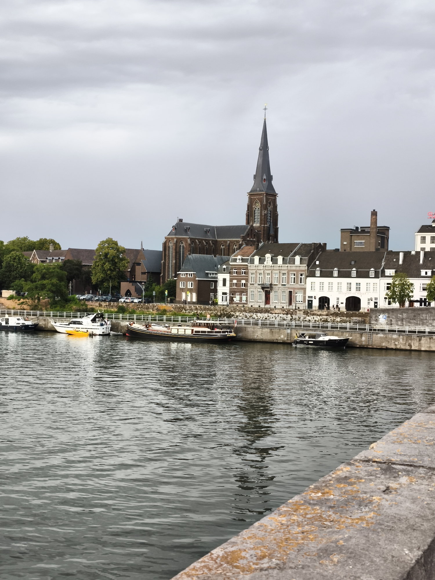 The image depicts a waterfront scene in Maastricht, Netherlands, focusing on the city's historic architecture and the Maas river. In the background, a large Gothic-style church with a prominent spired tower dominates the skyline. Adjacent to the church are several traditional European buildings with pitched roofs and multiple stories, some featuring arched windows and decorative facades. The buildings line the riverbank, separated from the water by a stone embankment and a white railing.
The Maas river flows in the foreground, its surface rippled by a gentle breeze. Several boats are moored along the embankment, including a long, dark-hulled barge, a white motor yacht with a yellow tender, and smaller white boats. The reflections of the buildings and the sky are visible on the water's surface.
The sky above is overcast with a mix of light and dark grey clouds, suggesting a mild or perhaps slightly cool day. The overall atmosphere is serene and picturesque, characteristic of many European river cities. The perspective is from a slightly elevated position, looking across the river towards the city. A stone ledge, with patches of lichen, occupies the lower right corner of the frame, suggesting the photo was taken from a bridge or a similar vantage point. A few people are visible walking along the embankment in front of the buildings, adding a sense of scale and life to the scene. The trees on the left side of the image suggest a green, natural element within the urban landscape.