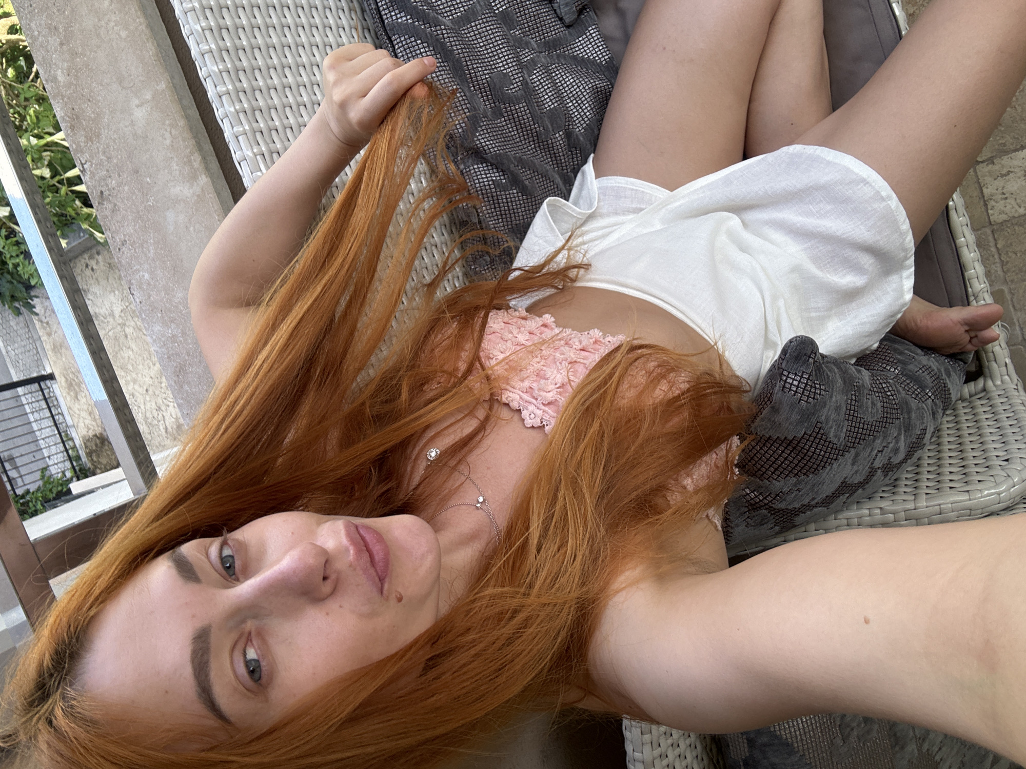 A woman with long, vibrant red hair is captured in a close-up, high-angle selfie while lying outdoors on a lounge chair, likely on a patio or balcony in Budva, Montenegro. She is looking directly at the camera with a neutral expression, her blue-gray eyes prominent. Her hair cascades around her, with one hand gently holding a portion of it near the top left of the frame.
She is wearing a light-colored outfit, including a pink, textured top and a white, loose-fitting garment that appears to be a dress or shorts. A delicate silver necklace with small, sparkling pendants adorns her neck. Her legs are visible, crossed and relaxed, resting on a dark grey pillow with a geometric pattern, which is placed on a light-colored, woven (wicker-style) outdoor chair. Another similar pillow is visible near her head.
The background shows elements of an outdoor setting: a concrete or stone wall, a metal railing, and lush green foliage, suggesting a pleasant, natural environment. The lighting is bright and natural, indicating it is daytime. There is no visible text in the image.