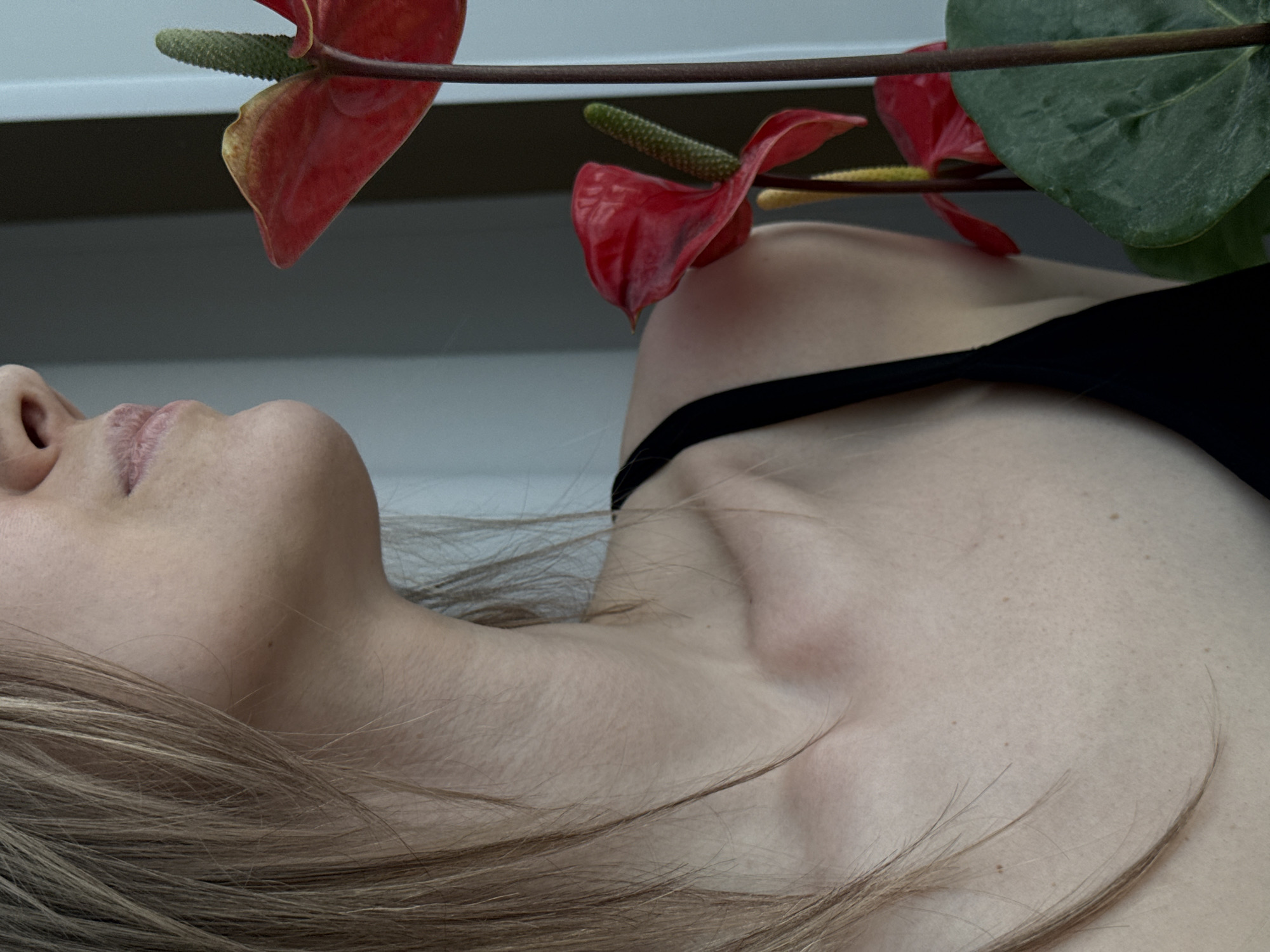 A close-up shot shows a woman lying on her back with her head tilted to the left. Her face is in profile, showing her nose and lips. She has light brown hair. She is wearing a black top. Three red anthurium flowers with green spadices are positioned above her head and neck. The background appears to be a light-colored surface. The city of Chaikovsky, Russia is not apparent in this image.