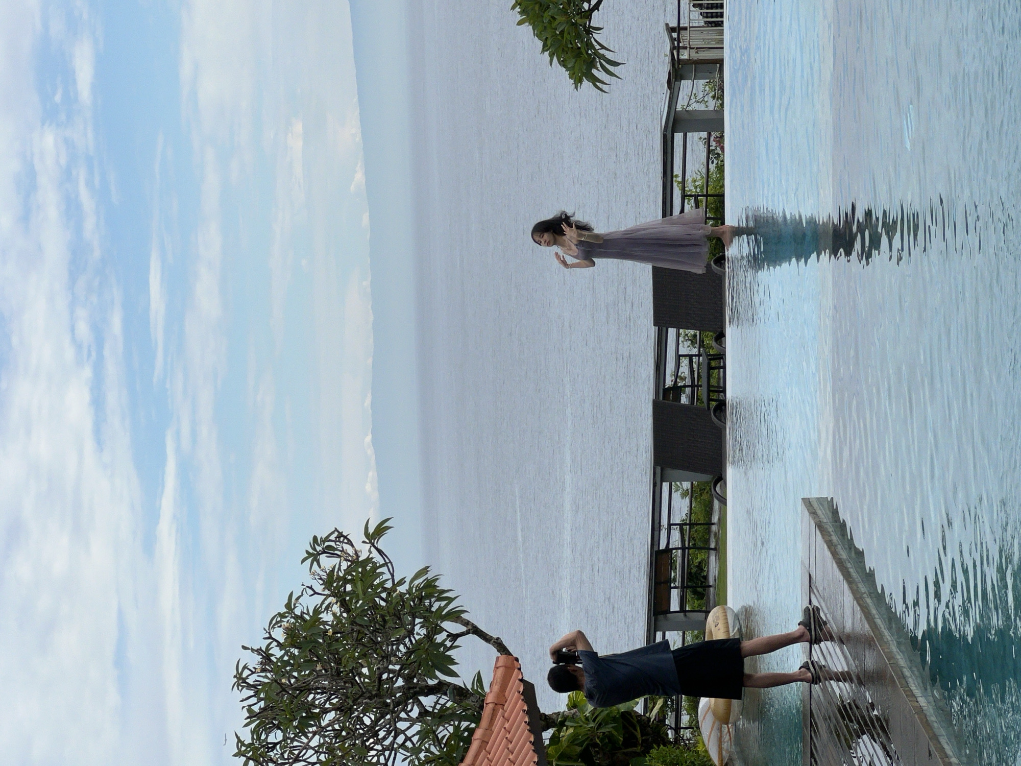 This image depicts an outdoor scene at a scenic location, likely a resort in Kuta Selatan, Indonesia, during bright daylight hours. The primary subjects are two people engaged in a photography session overlooking a vast body of water.
In the mid-ground, a woman with long dark hair is seen standing gracefully at the edge of an infinity pool. She is wearing a light-colored, long dress and has her arms slightly raised, appearing to pose for a photograph. Her gaze is directed towards the expansive, calm ocean or large lake that stretches to the horizon in the background.
In the lower-left foreground, a man, presumably a photographer, is captured from behind. He is dressed in a dark t-shirt and shorts, bending down with a camera to his eye, focusing on taking a picture of the woman. He stands on a wooden deck adjacent to the infinity pool. A partially visible yellow inflatable ring rests near his feet.
The infinity pool occupies the right side of the frame, reflecting the sky and showing subtle ripples on its surface. Beyond the pool and the woman, the large body of water appears slightly hazy on the horizon, meeting a light blue sky dotted with scattered, wispy white clouds. Tropical foliage, including a tree with green leaves and a glimpse of a terracotta-tiled roof, is visible on the left, adding to the lush, resort-like atmosphere. The scene conveys a relaxed and picturesque setting for leisure and photography. There is no visible text in the image.
