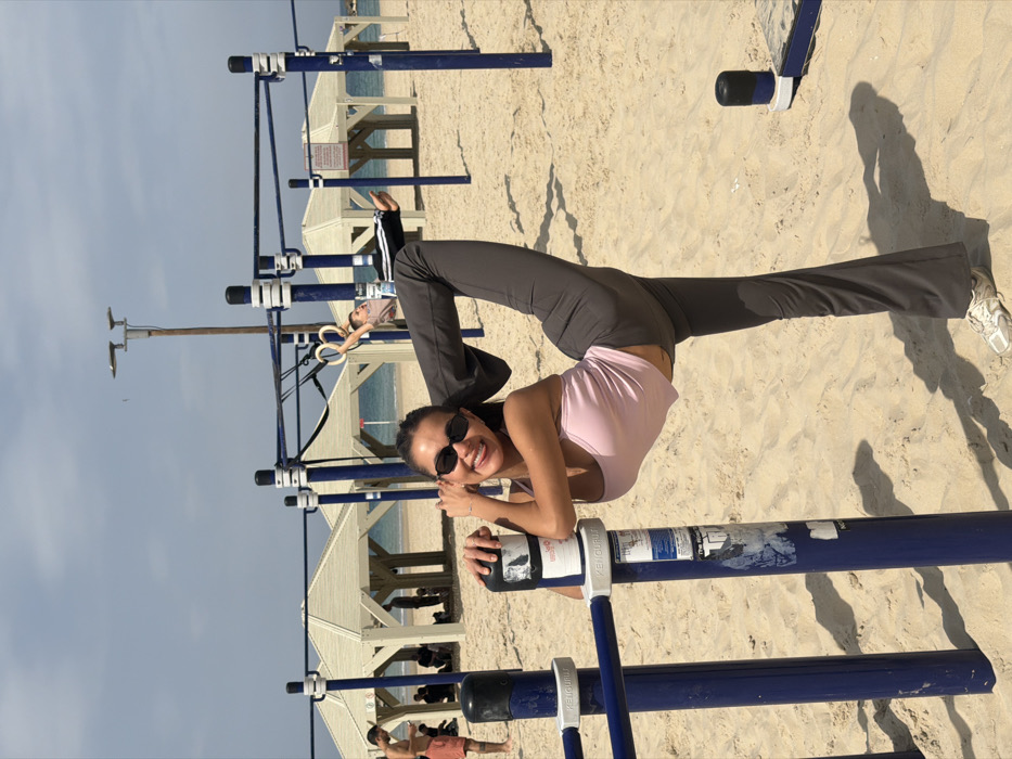 The scene depicts a public beach calisthenics area in Tel-Aviv, Israel. A female, positioned centrally, performs a high leg raise with her right leg extended vertically. She wears small dark sunglasses, a light pink tank top, and dark grey athletic pants, with white athletic shoes on her left foot. Her right hand grips a dark blue vertical metal pole, part of the exercise equipment, which displays several partially peeled white labels, including text like "KENGURUT" and "TRI." Her left hand is bent near her face, and she smiles towards the camera.
In the midground, another male individual hangs from gymnastic rings attached to an adjacent section of blue and white metal calisthenics bars, performing a horizontal body hold. Further into the background, several white, hexagonal-roofed sunshades are visible on the sandy beach, with people seated beneath them. The Mediterranean Sea forms the horizon, under a clear sky with scattered clouds. A tall utility pole with two lamps stands in the mid-background. Shadows are distinctly cast on the sand.