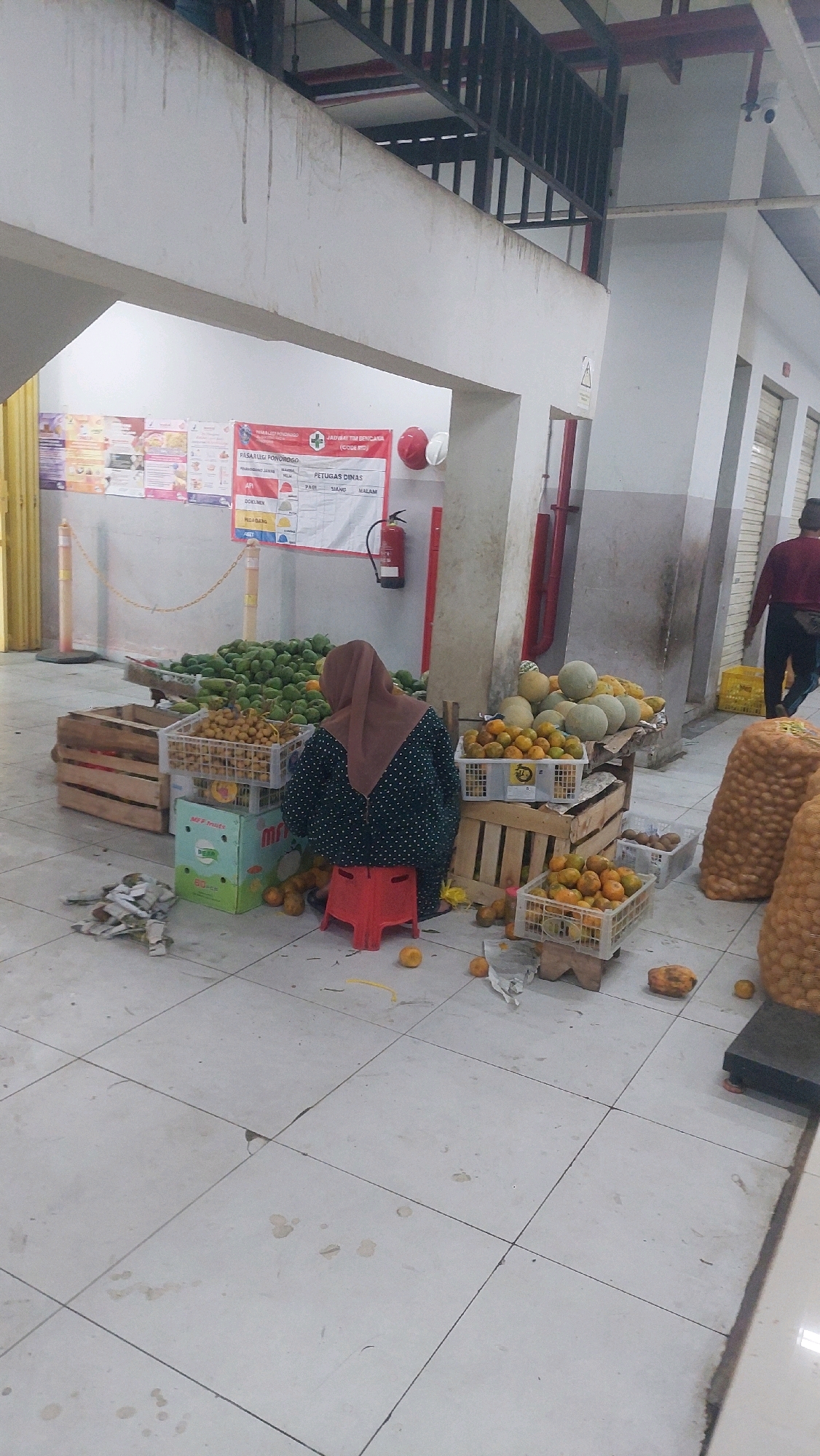 A person wearing a hijab and a dark polka-dot patterned jacket is sitting on a red stool in front of a fruit stall in what appears to be an indoor market. The stall displays various fruits including green papayas, oranges, and melons. Some of the fruit is in crates, while other produce is piled up. There are also sacks of produce visible in the background. The floor is tiled and appears somewhat dirty with some spills. In the background, there is a fire extinguisher mounted on a wall and a sign with text in Indonesian. Another person in a burgundy shirt and dark pants is walking away from the stall towards the right side of the frame. The environment suggests a local market or a similar commercial space in Ponorogo, Indonesia.