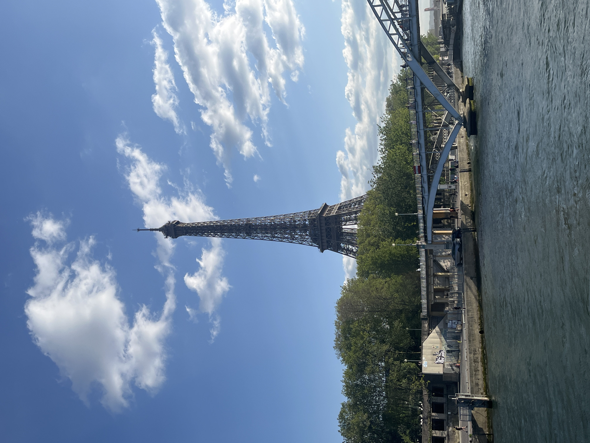 This full-shot photograph captures a vibrant daytime view of the Eiffel Tower in Paris, France, positioned prominently on the left. The scene is bathed in bright sunlight under a clear blue sky, adorned with scattered, fluffy white cumulus clouds, indicating fair weather, likely during midday or early afternoon.
The iconic Eiffel Tower stands tall and majestic, its intricate metalwork silhouetted against the bright sky. To the right, the wide, greenish-blue waters of the River Seine flow horizontally across the frame. A large, grey metal bridge, characterized by its robust trusses and girders, occupies a significant portion of the foreground and midground on the right, appearing to span the river. The perspective suggests the photo was taken from a vantage point along or near this bridge, looking towards the tower.
Along the riverbanks, a line of dense green trees provides a natural border. Below the trees, concrete walls, walkways, and some small buildings are visible. Faint, illegible graffiti or markings can be observed on some of these riverside structures. Several people are discernible both on the bridge and along the riverbank, engaged in walking or observing the surroundings. Lampposts are also scattered along the river's edge. The overall impression is a classic, sunny Parisian landscape.