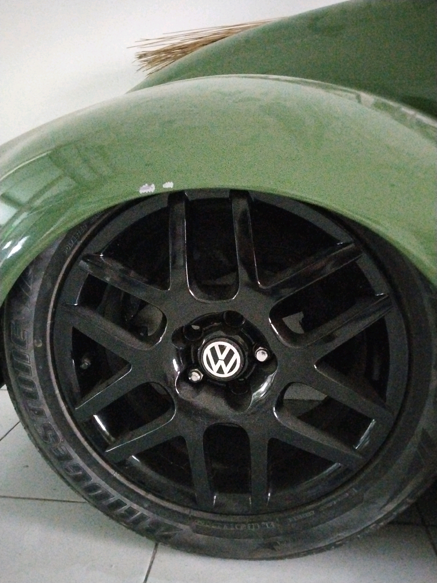 A close-up, low-angle shot shows the front wheel of a green Volkswagen Beetle with aftermarket black alloy rims. The car's fender is visible above the wheel, and a broom made of dried grass is leaning against the car's bodywork in the upper portion of the frame. The scene is set indoors, likely in a garage or workshop, with a tiled floor visible beneath the car. The overall lighting is dim, with a slight green tint to the image. There are no people present in the image. The image does not provide any information about its specific location in Sinduadi, Indonesia.