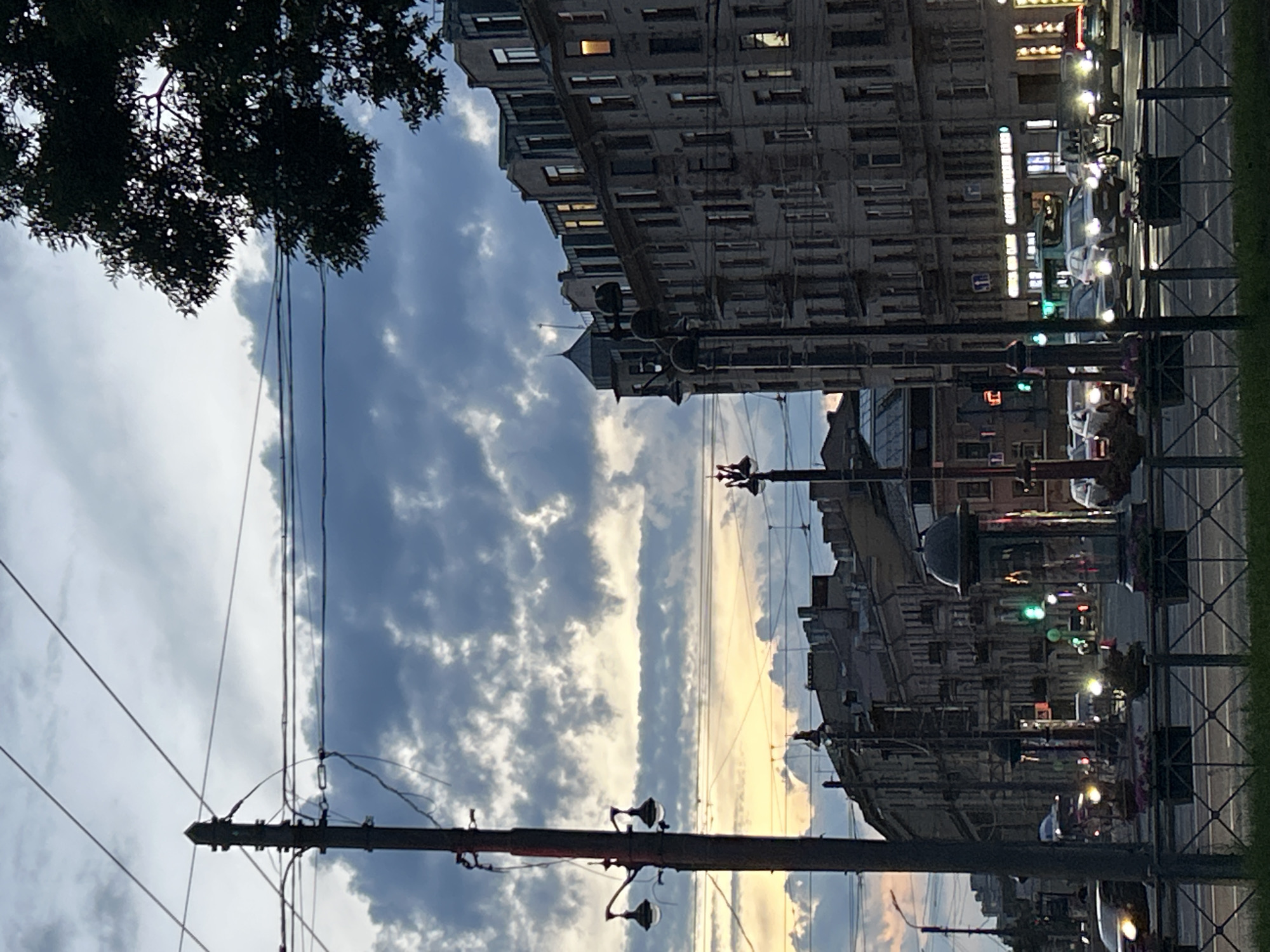 The image depicts a street scene in a city at dusk. The sky is filled with dark, cloudy formations, with hints of yellow and orange light near the horizon. In the foreground, a dark pole with wires stretches vertically, supporting streetlights and electrical equipment. To the right, a tall, multi-story building with a mansard roof and numerous windows dominates the cityscape. Some windows are illuminated, suggesting interior lights. Further down the street, other buildings with similar architectural styles are visible, along with more streetlights and utility poles. Traffic is present on the street, with cars and buses visible, some with their headlights on. Traffic lights can be seen, one of which is green. Planters with flowers are placed along the side of the road. The overall atmosphere is that of a city transitioning from day to night, with a sense of urban activity.