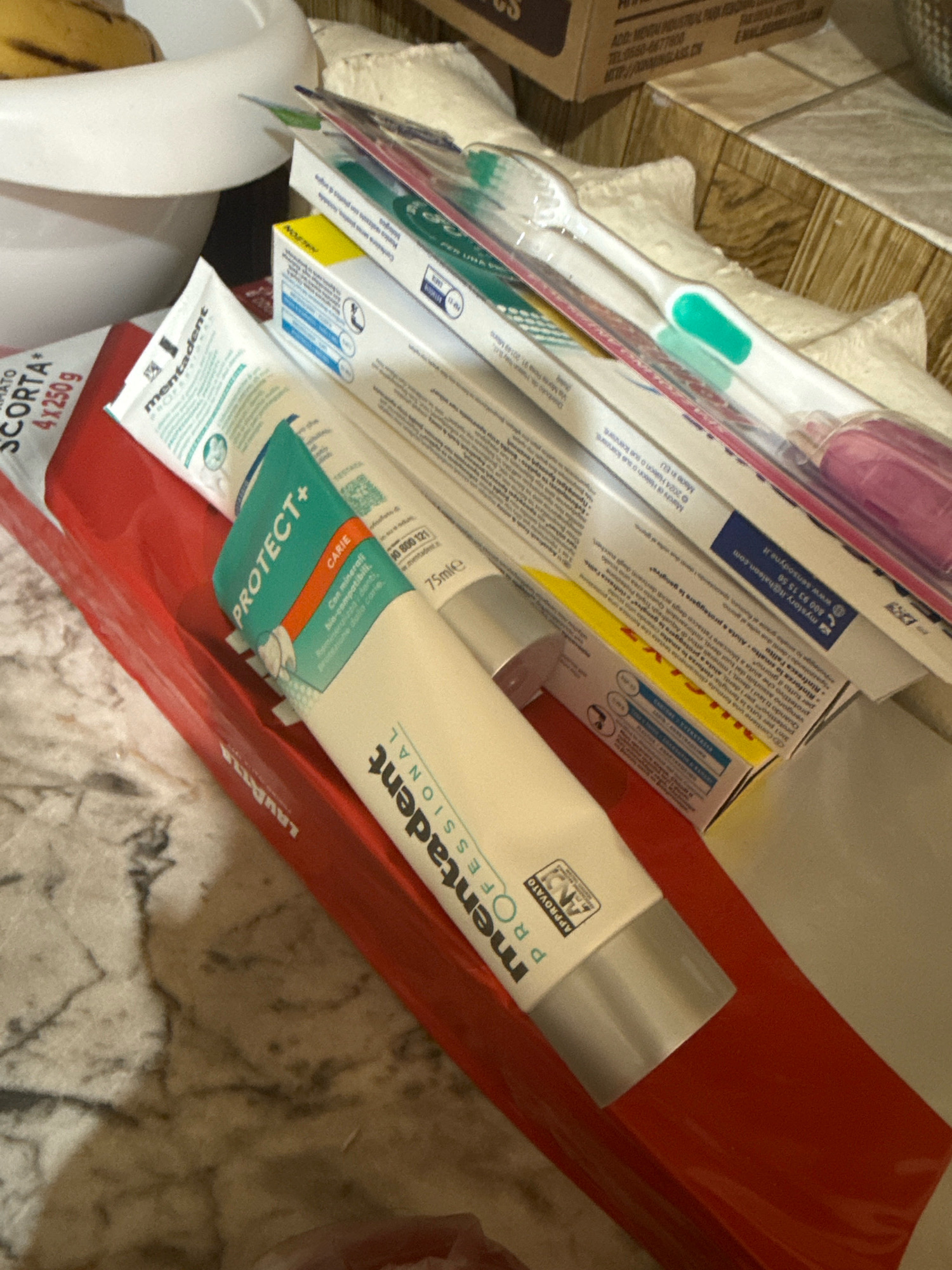 The image shows a close-up, top-down view of a collection of personal hygiene products, specifically toothpastes and toothbrushes, arranged on a marbled countertop. A white ceramic bowl containing what appears to be bananas is visible in the upper left corner. Several tubes of toothpaste, predominantly white with green and orange accents, are prominent. One tube clearly displays "mentadent PROTECT+ CARIE" in green lettering. Another tube, also white, has "mentadent PROFESSIONAL" printed on it in black text. Several boxes of toothpaste are stacked behind the tubes, with visible text including "made in EU." A pink-handled toothbrush is also visible, along with other toothbrushes with green accents. A red rectangular object, possibly a box or packaging, lies beneath the toothpaste tubes, with the word "LAVAZZA" partially visible in white lettering. The overall environment suggests a domestic setting, likely a bathroom or kitchen counter. There are no people or any indication of the city of Basarabeasca, Moldova.