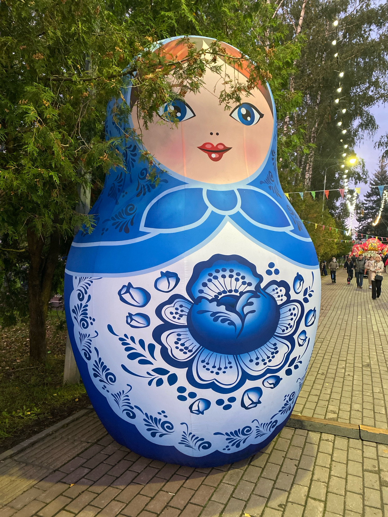 An inflatable Matryoshka doll, approximately 20 feet tall, is displayed outdoors. The doll is painted with traditional Gzhel-style blue and white floral patterns and features a face with blue eyes, pink cheeks, and red lips. It stands on a brick-paved walkway lined with trees and string lights. In the background, people are walking and riding scooters. The scene appears to be in a park or public square, likely during an evening event given the lighting. The location is identified as Ivanteyevka, Russia.
