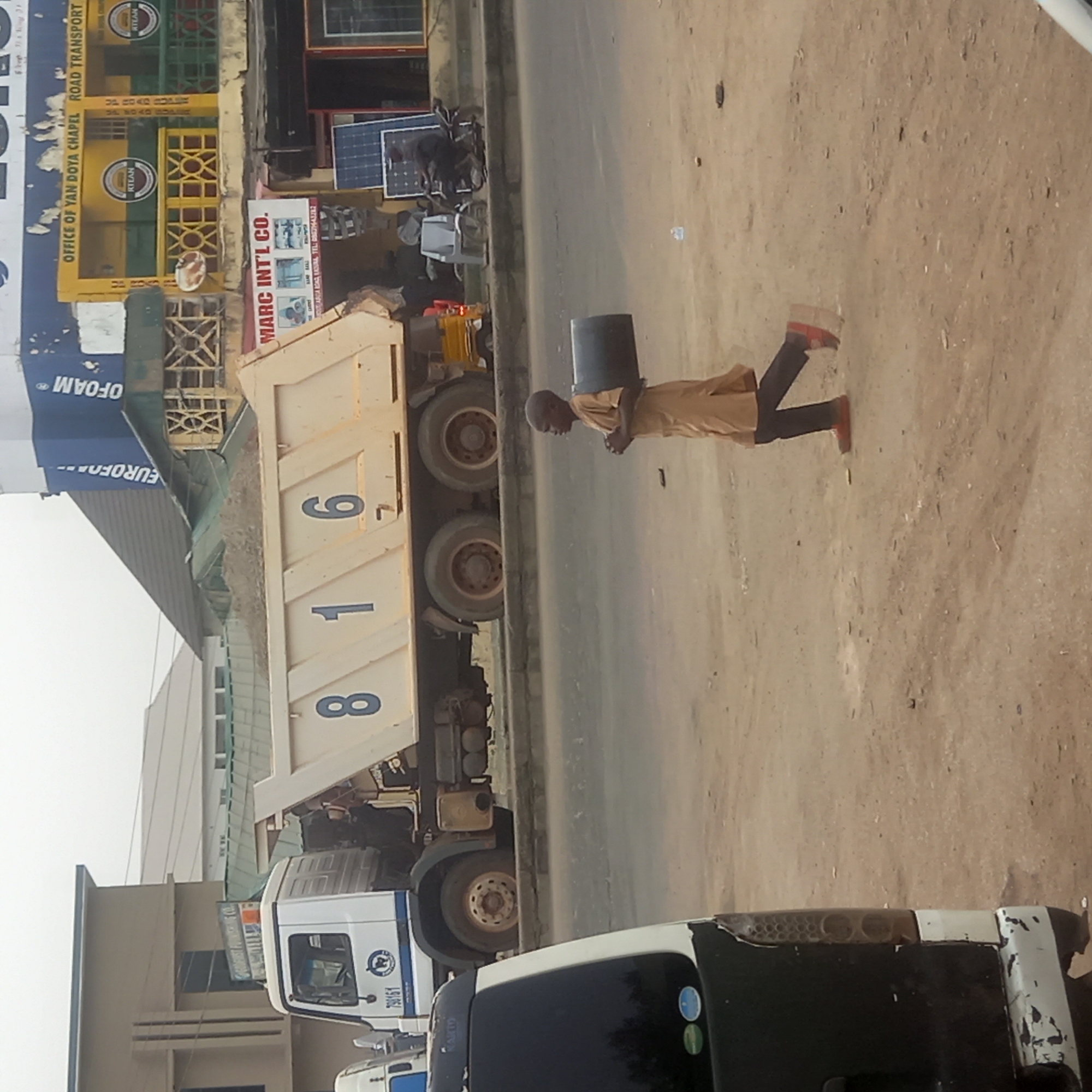 This image captures a street scene in Dadi Riba, Nigeria, on what appears to be a bright, sunny day. The primary subject is a person, likely a young man, walking on the right side of a dirt road or shoulder, carrying a dark bucket balanced on their head. They are wearing a light brown or tan long-sleeved outfit and dark trousers with reddish-brown shoes.
To the left of the person is a large, light-colored dump truck, laden with what looks like sand or gravel. The numbers "816" are visible in dark blue on the side of its bed. Further left, part of a white truck cab is visible, with "798161" printed on its side and a blue circular logo featuring a globe. In the foreground, the rear of another vehicle, possibly a white van with a dark tinted window, is partially visible.
Along the left side of the road are several commercial buildings or shops. One building features a blue and white sign with the partially visible text "EUROFO---" and "OFOAM". Another building has a prominent yellow and green sign with "OFFICE OF YAN DOYA CHAPEL ROAD TRANSPORT" and "RITEAN" logos. Below this, a white sign for "MARC INT'L CO." is visible, with smaller text that includes "АЙСА КАД BARA, TEL:ЕН43282" and mentions "UP ROAD". Solar panels are displayed outside one of the shops. The overall setting is an active commercial roadside.