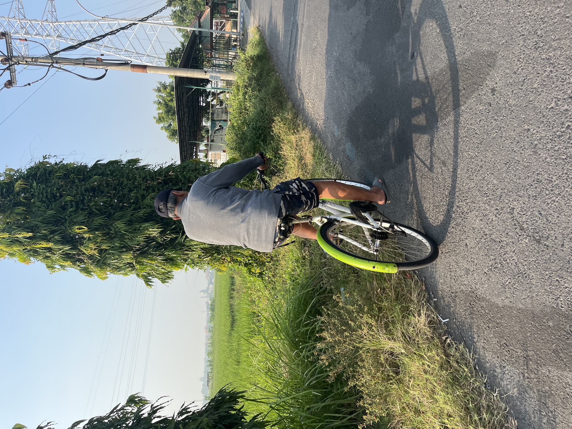 A person is riding a bicycle down a paved road, seen from behind. The person is wearing a gray long-sleeved shirt, dark patterned shorts, a black baseball cap, and sandals. The bicycle is white with a bright green fender over the rear wheel. The road has a visible shadow cast by the cyclist and bicycle. To the left of the road is a field of green grass and then a vast rice paddy. To the right of the road, there is a tall, thin tree and further back, a utility pole with electrical wires and a large metal tower. Behind the utility pole, there are several simple structures with roofs, possibly shelters or small buildings. The sky is clear and light blue. The overall impression is a rural or semi-rural setting. The image is taken outdoors in what appears to be a tropical climate, likely in Indonesia.