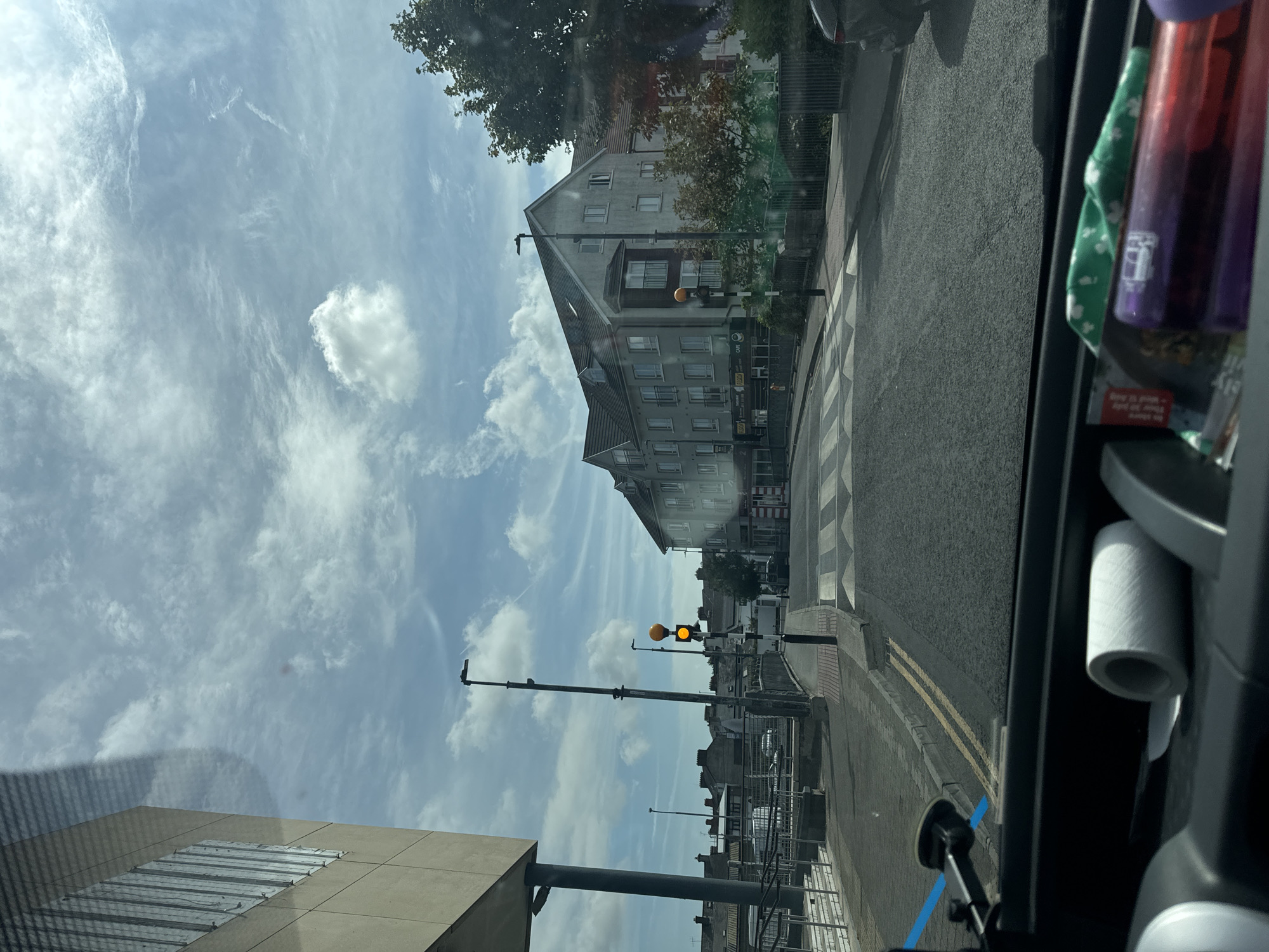 The image captures a view from a vehicle looking out towards a street scene in Waterford, Ireland. The perspective is from the driver's seat, with the dashboard and windshield occupying the foreground. On the dashboard, a roll of white material, possibly toilet paper, and a few other objects are visible, including a green patterned cloth and a purple cylindrical object.
The scene outside the vehicle is dominated by a street with a pedestrian crossing marked by white stripes. A traffic light is visible, displaying a yellow signal. On the left side of the street, a modern, light-colored building with vertical window elements stands out. Further down the street, a multi-story building with apartments or offices is visible, featuring multiple windows and balconies. Trees and other foliage are present along the side of the road. The sky is partly cloudy with a blue sky peeking through. The road surface is asphalt, and there are yellow double lines indicating traffic lanes. In the background, several other buildings with pitched roofs are visible, suggesting a mix of architectural styles. The overall impression is a typical urban street scene on a daytime.
