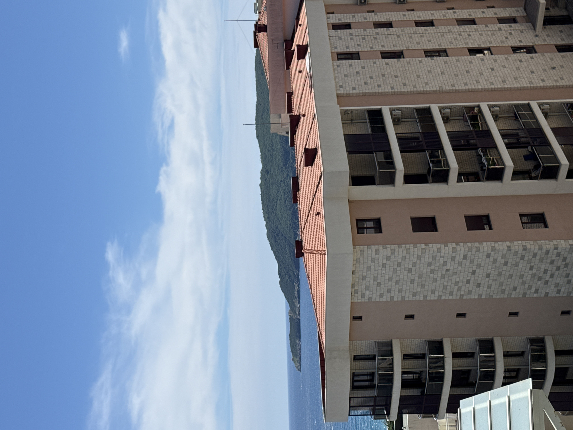 This is a photograph of a building with a tiled roof, situated on a coastline with a lush green hill rising behind it and a body of water in the foreground. The sky is a clear blue with some wispy white clouds. The building itself appears to be multi-story with numerous windows. Some of the windows are open, revealing air conditioning units and what appear to be indoor items. The building's facade is a mix of light brown and grey tiled sections. A red-tiled roof with visible dark structures extending from it runs along one side of the building, sloping down towards the water.
The scene suggests a sunny day, likely in a Mediterranean climate given the greenery and the architectural style. The presence of air conditioning units indicates it's a warm season. The orientation of the image is sideways, making the building appear to be on its side. The location context indicates this is Boreti, Montenegro. There is no visible text in the image. The overall impression is one of a coastal resort or residential building in a scenic natural setting.