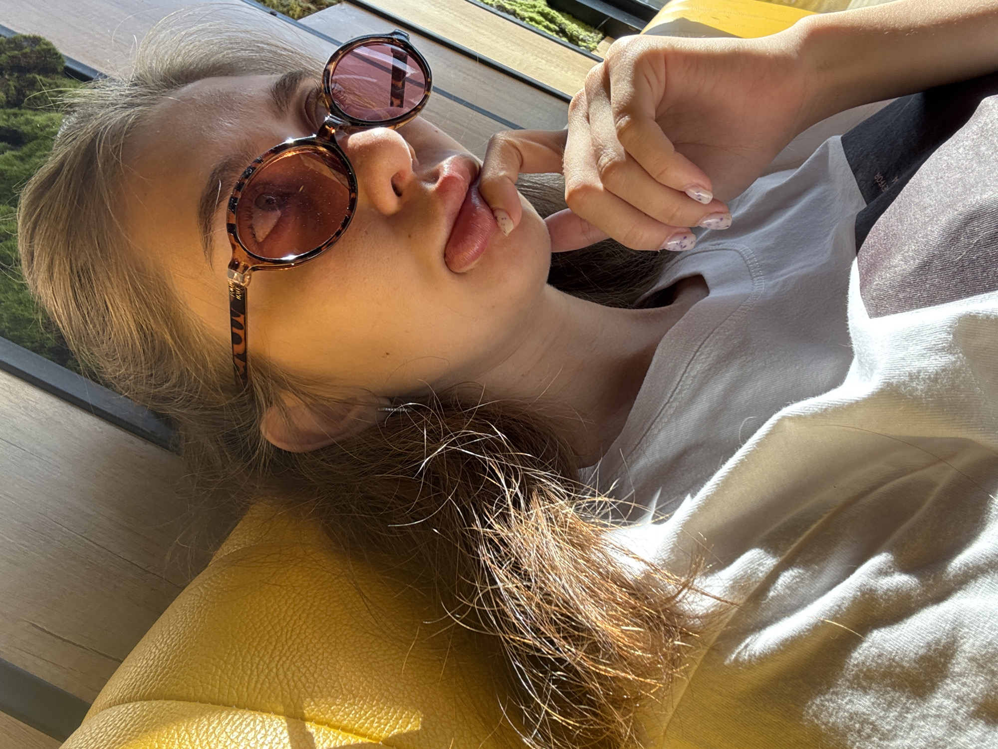 A young woman with light brown hair is pictured from the chest up, leaning back against a yellow textured surface that appears to be a chair or booth. She is wearing a white t-shirt and stylish tortoise-shell sunglasses with pink lenses. Her right hand is raised to her face, with her index finger touching her chin, and her mouth is puckered as if she is blowing a kiss or making a funny face. Her fingernails have a light-colored polish with subtle purple accents. The background shows wooden slats and greenery, suggesting an outdoor or well-lit indoor setting. The lighting indicates it's daytime, with bright sunlight casting shadows.