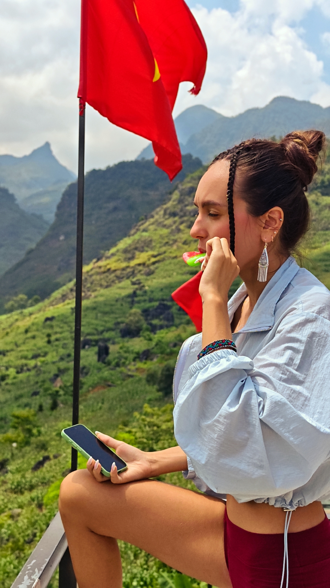 A young woman is sitting on a railing, eating a lollipop, and holding a smartphone, with a Vietnamese flag and mountainous scenery in the background. The woman has her hair styled in a bun with braids, wears dangling earrings, a light blue jacket, and red shorts. She is holding a smartphone with a green case in her left hand. The Vietnamese flag waves in the upper left corner. Behind her is a lush, green landscape with distant mountains under a partly cloudy sky. The photo was taken in Lũng Phìn Commune, Vietnam.