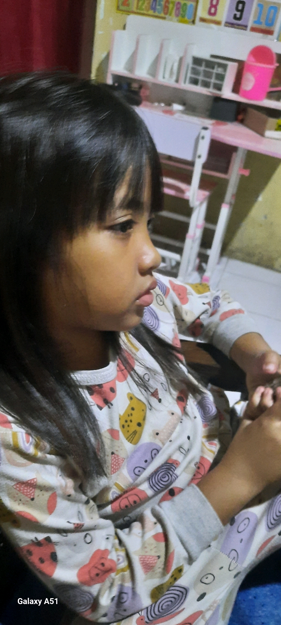 The photograph, taken in the city of Ploso, Indonesia, shows a young girl from the chest up, facing to the right. She has dark hair and is wearing a long-sleeved shirt with a pattern of cartoon animals. Her right hand is visible near her chest, and her left hand is not clearly visible. In the background, a pink desk and chair set is partially visible, along with a shelf unit holding various items. The lighting suggests an indoor setting, possibly a room in a house. The bottom left corner of the image displays "Galaxy A51," indicating the device used to capture the photo.